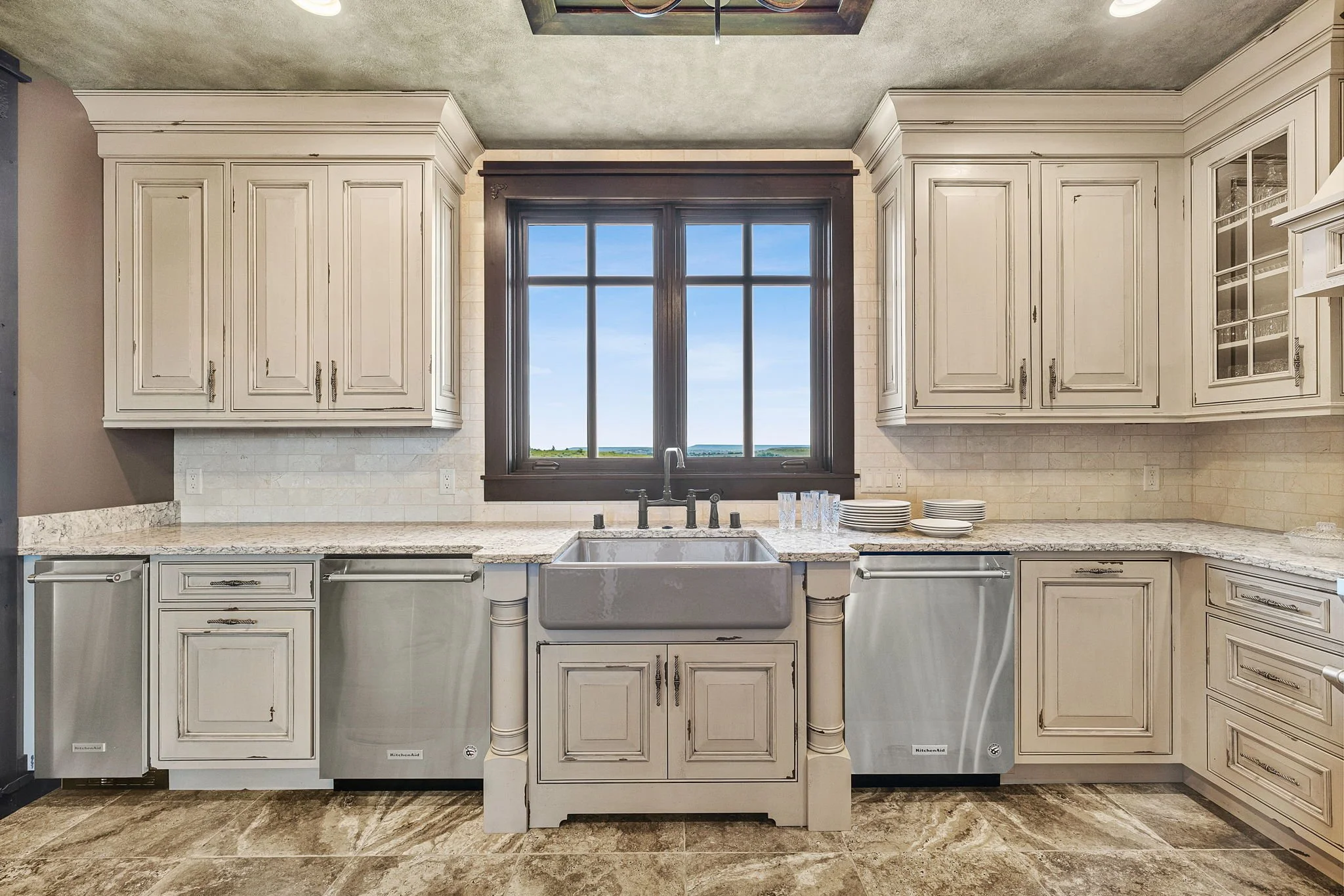 Kitchen with cream-colored cabinets, a large window with dark trim, and a farmhouse sink. The countertop has a marble pattern, and there are dishes stacked on the right side. The floor has a tile pattern, and the ceiling is painted in a muted green s