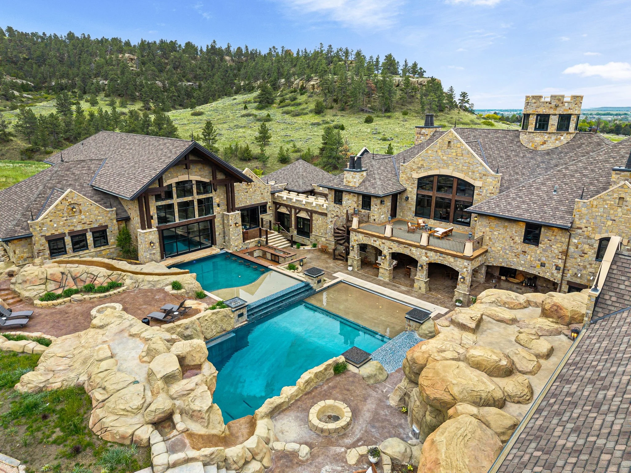 A luxurious mansion with stone and wood exterior, multiple rooftops, a swimming pool, and outdoor leisure areas. The property features large windows, a hot tub, and scenic mountain views in the background.