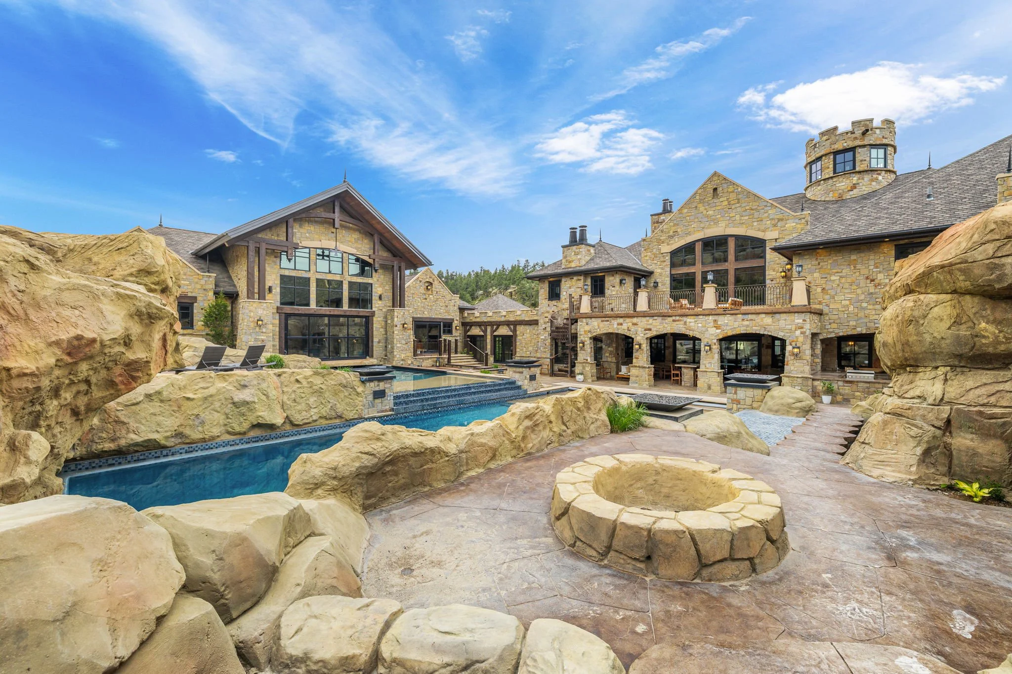 Large stone mansion with multiple levels, large windows, and stone walls. The backyard features a rocky landscape with a swimming pool, hot tub, and firepit, under a blue sky with scattered clouds.