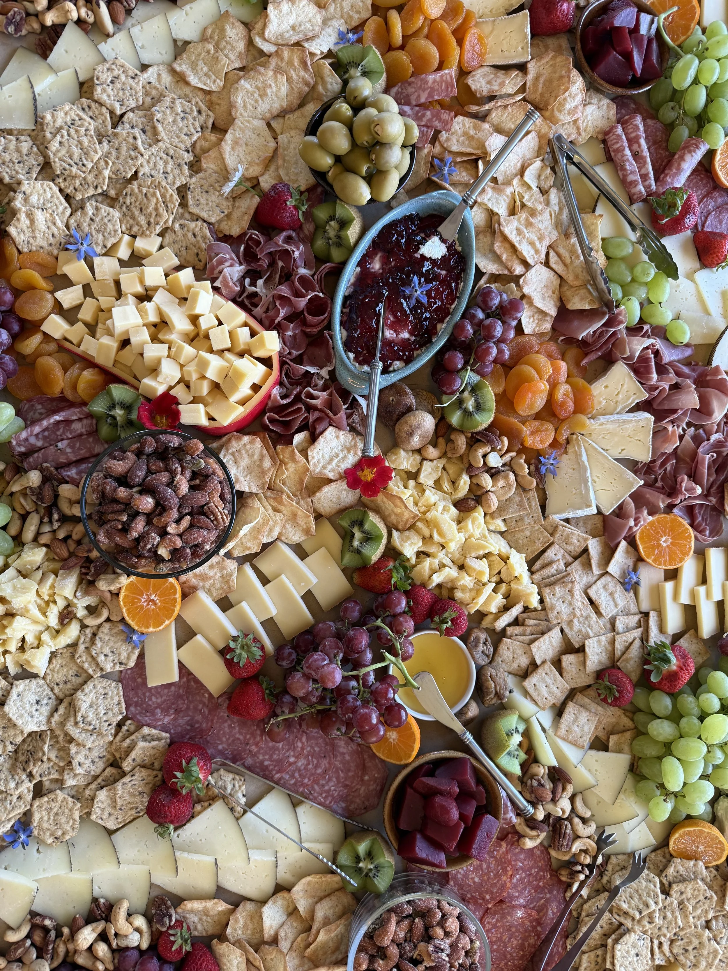 A close-up view of a charcuterie board that Heather Leith made for a wedding reception featuring sliced cheeses, crackers, grapes, strawberries, various meats, fruit preserves, nuts, and decorative edible flowers.
