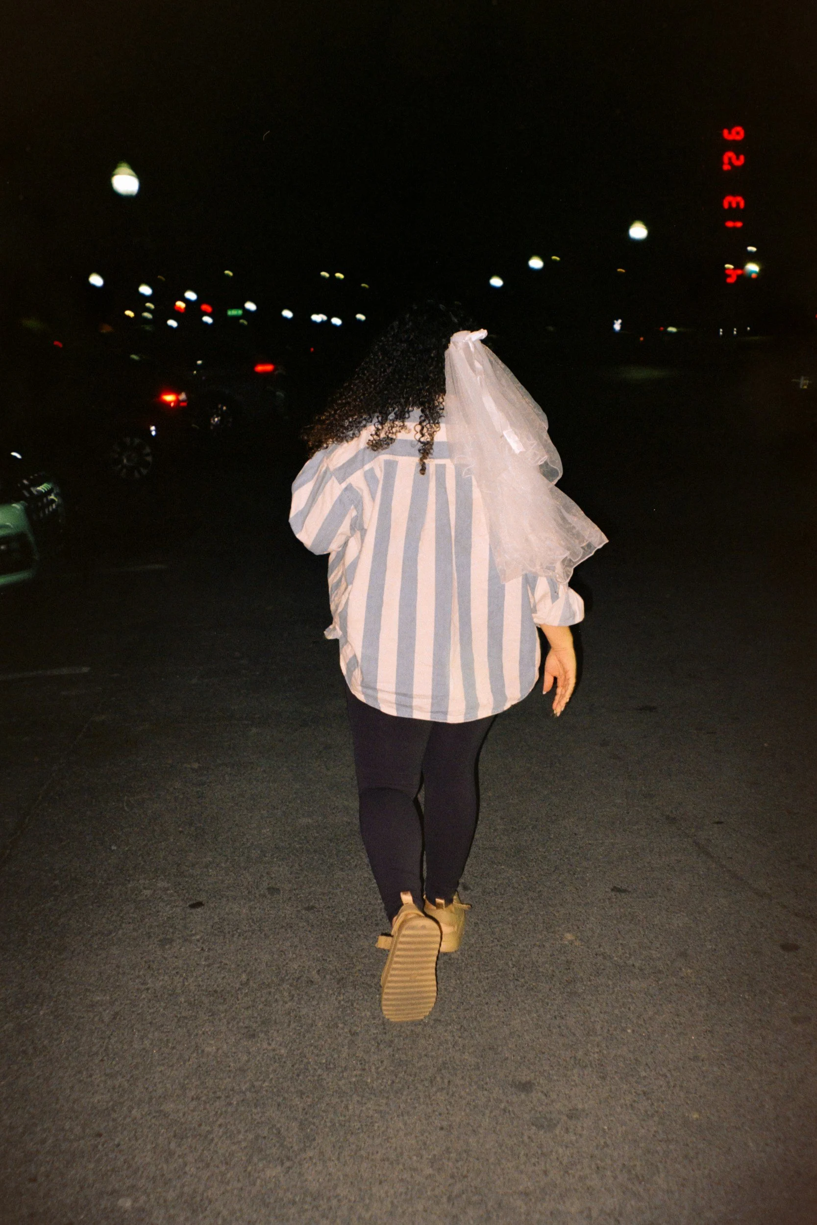 A flash photo of a casually dressed bride at her bachelorette party in a striped shirt, black leggings, and a veil walking on a dark street at night.