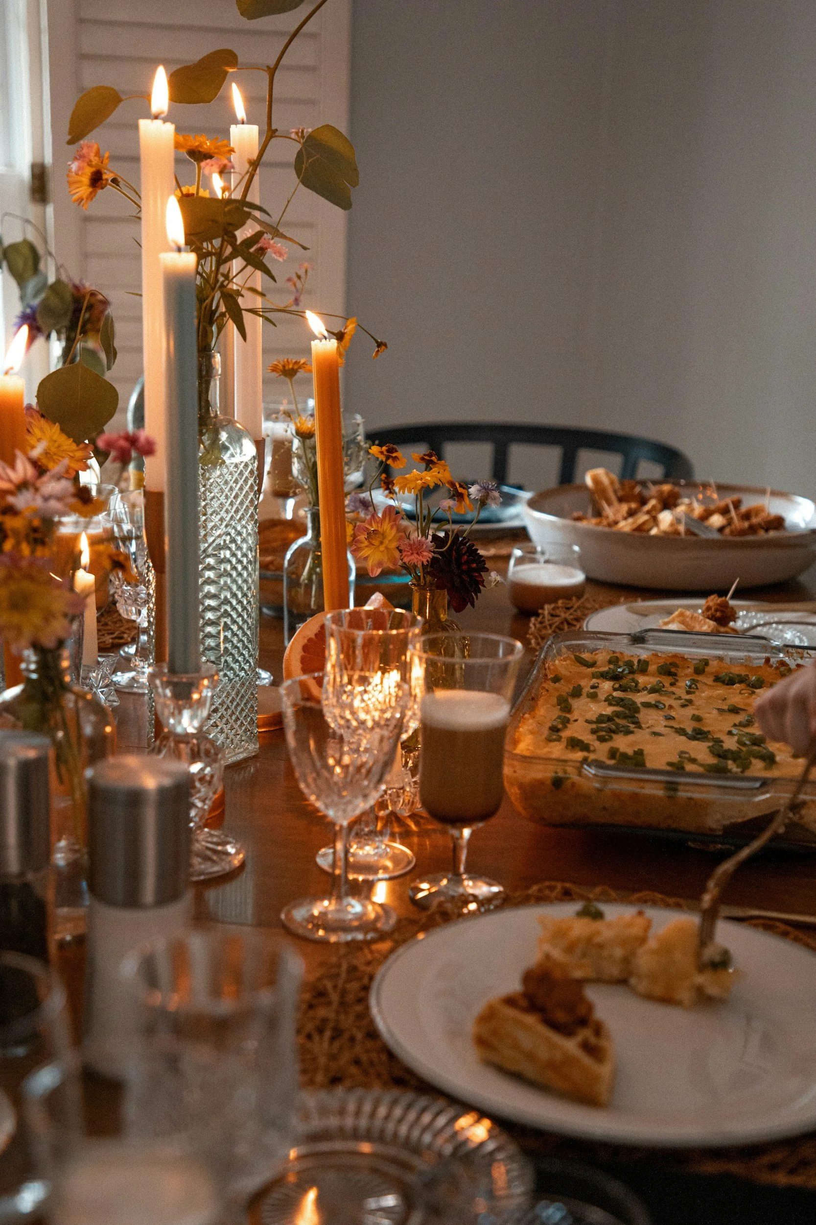 A fall-themed dining table decorated with lit candles, flowers in glass vases, and glasses of drinks. There are plates with food including a rectangular dish of casserole with green herbs and a plate with pieces of pie.
