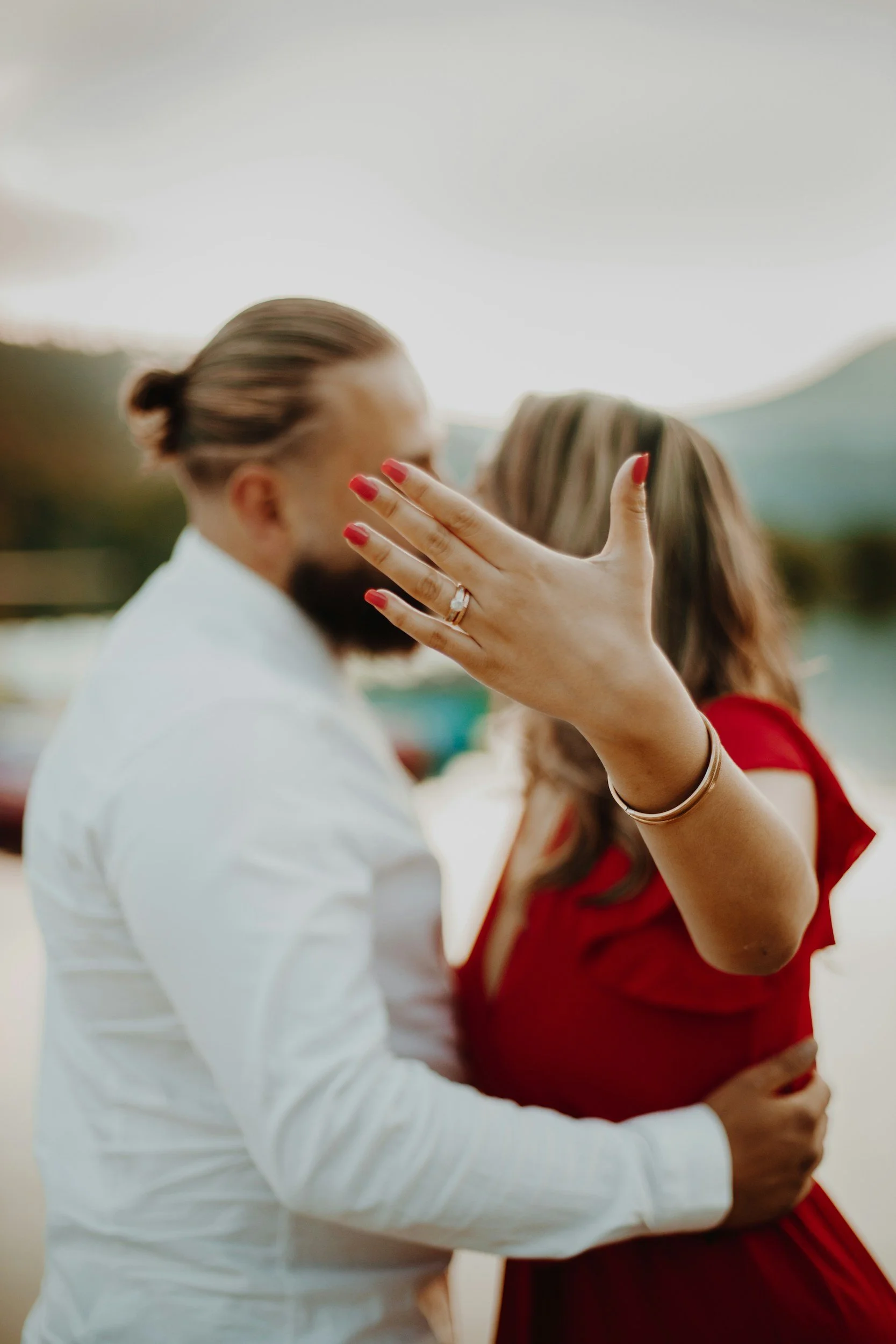 A couple kissing, with the woman flashing her engagement ring on her left hand. The woman is wearing a red dress, and the man is in a white shirt. The focus is on the woman's hand and ring, with the couple's faces slightly blurred.