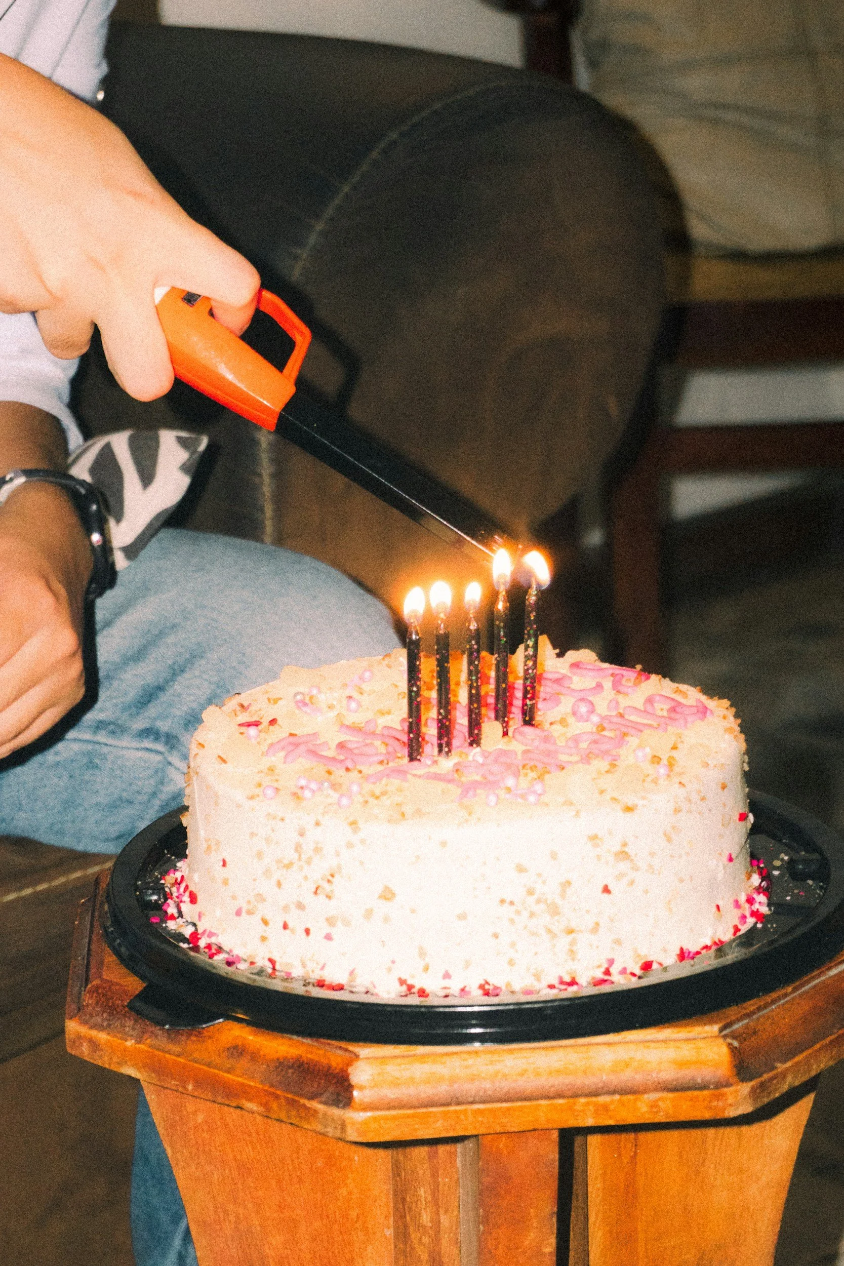 A person is holding a lighter over a round, decorated birthday cake with pink and white icing and sprinkles, placed on a wooden stand.