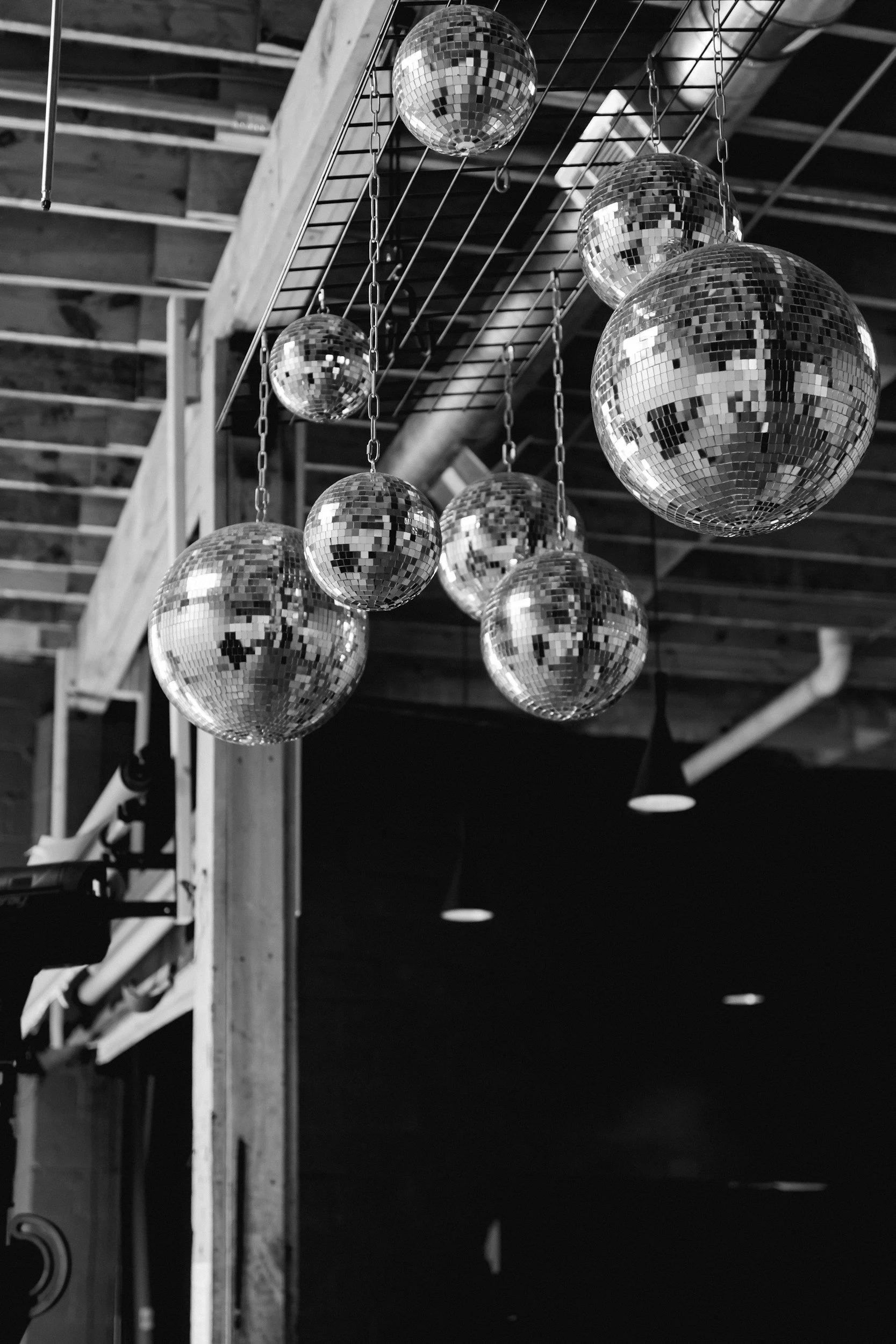 Multiple disco balls hanging from chains, reflecting light, in loft-style interior with exposed wooden beams and ceiling pipes.