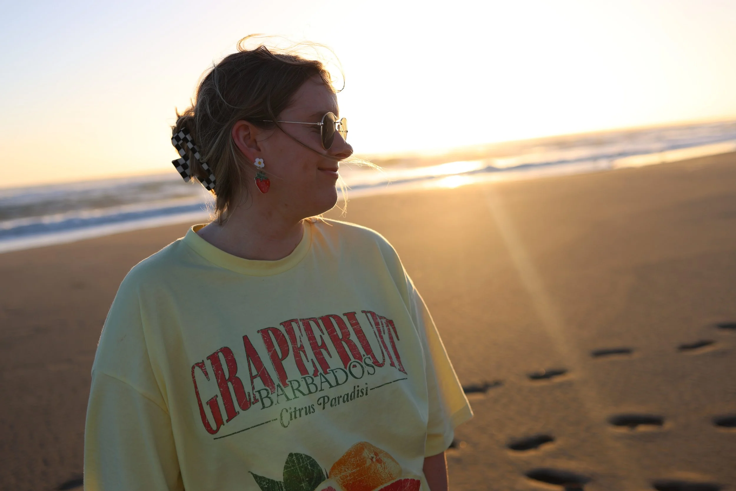 Heather Leith, a freelance copywriter for weddings and events, stands on a beach during sunset wearing sunglasses and strawberry earrings. Her yellow shirt says "Grapefruit."