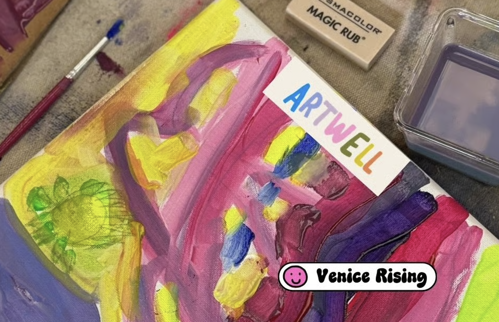 Close-up of an abstract acrylic painting with bright colors including yellow, pink, purple, and green, with a sticker labeled 'ARTWELL' and a sign that says 'Venice Rising' with a smiling face icon.