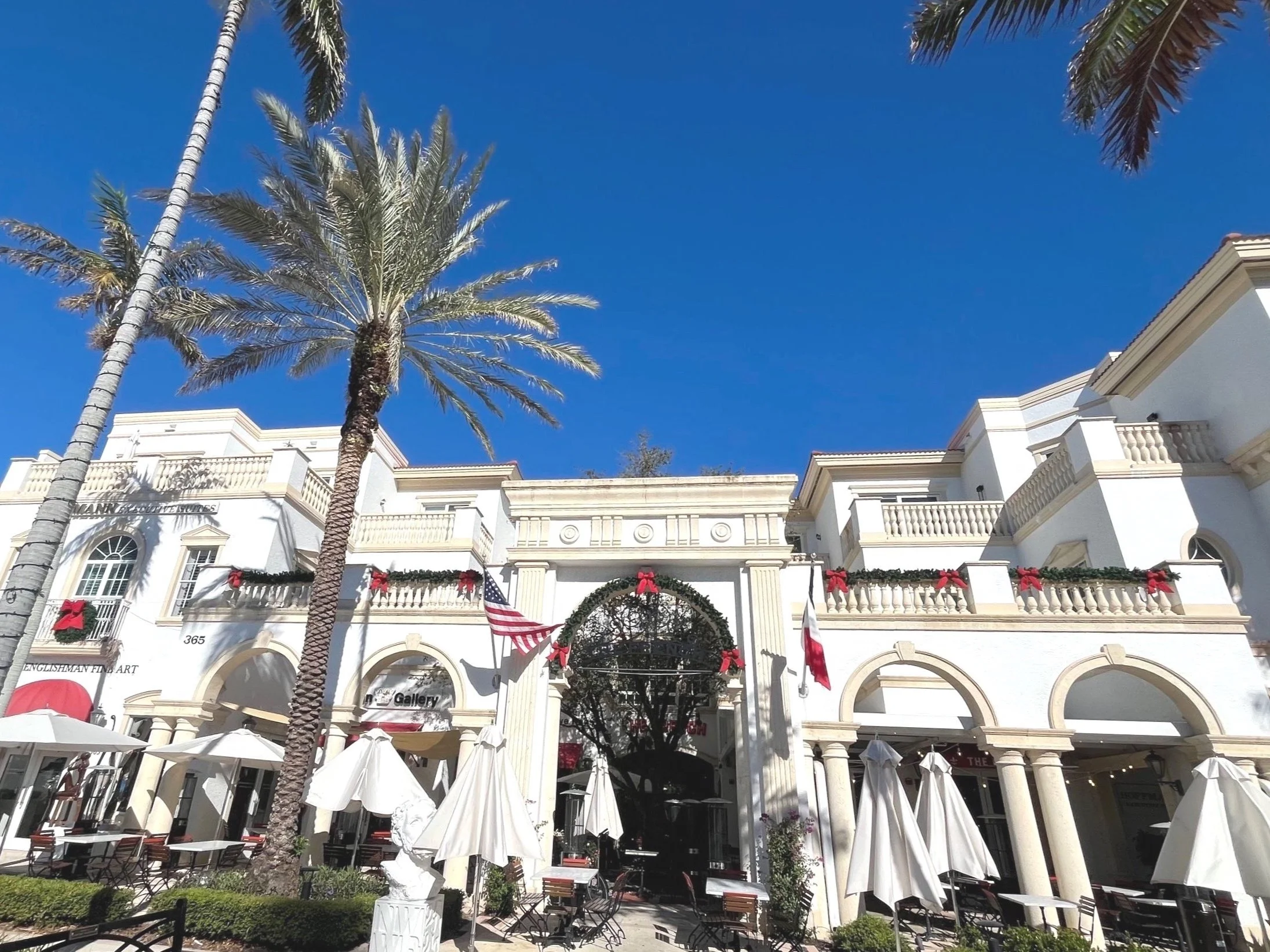 Colonial-style building with balconies and arches, adorned with holiday decorations, palm trees, and outdoor seating area under umbrellas against a blue sky.