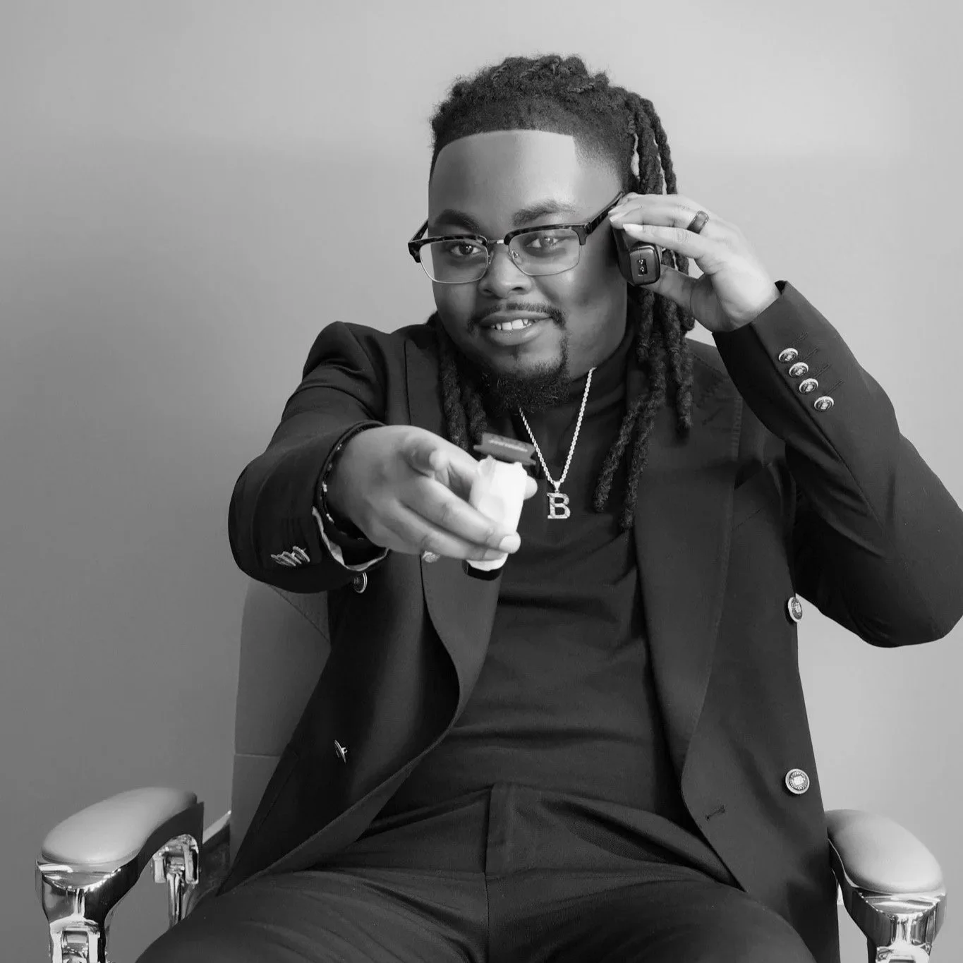 A man with glasses and dreadlocks sitting in a chair, smiling. He wears a suit with a pendant necklace and holds a remote control while talking on a phone.
