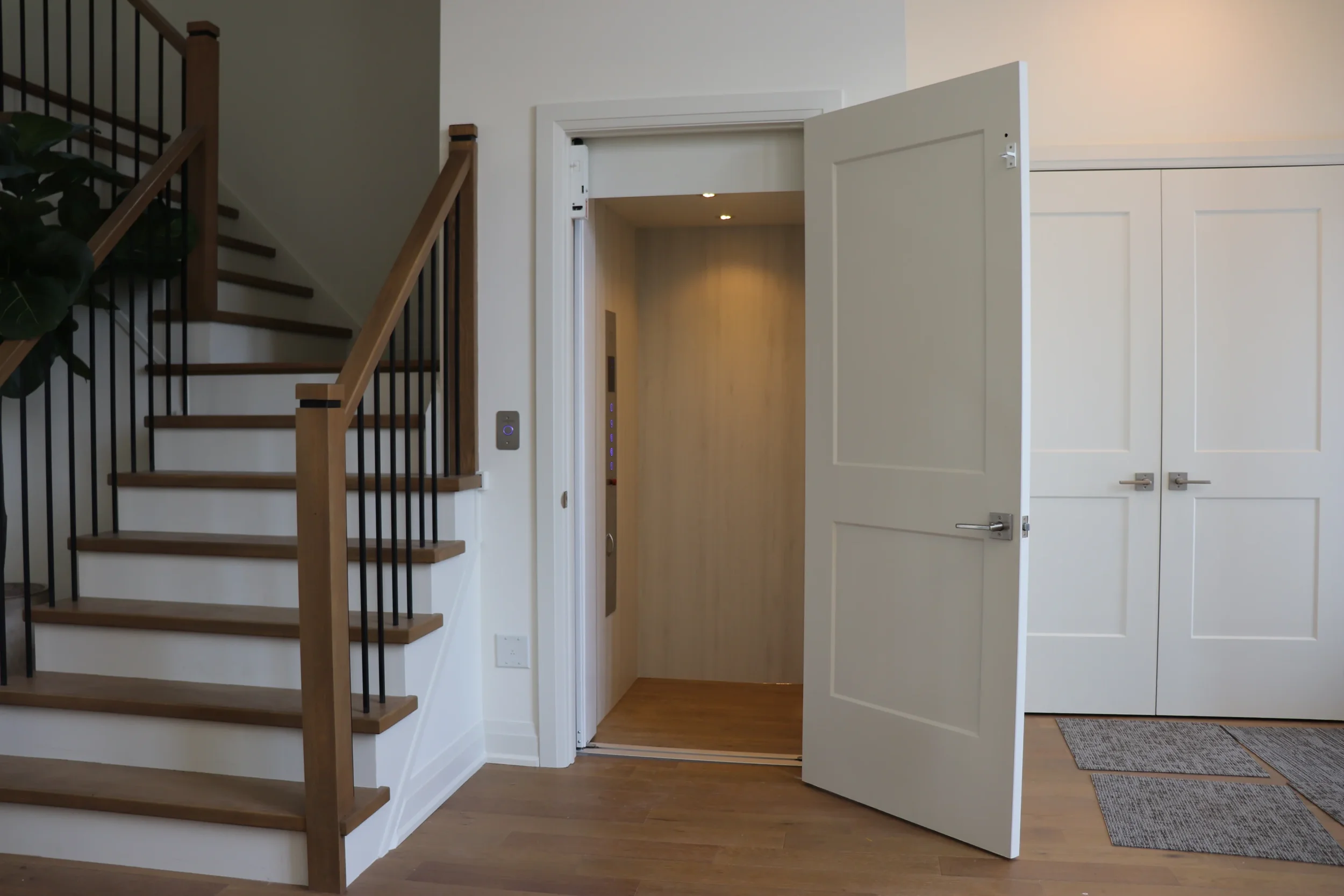An interior view of a residential space featuring a staircase with wooden steps and black metal balusters on the left, an open door leading to an elevator with wood paneling and LED indicators on the right, and a large double closet with white paneled doors in the background.