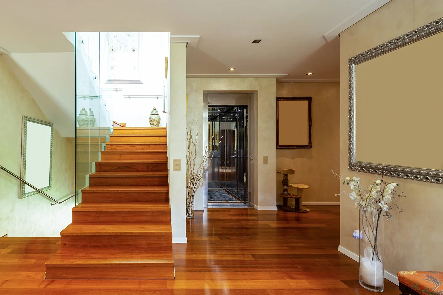 Interior view of a home with wooden flooring, a staircase with wooden steps and glass railing, and decorative vases on top of the staircase. An elevator with mirrored doors is visible, along with artwork frames and decorative plants.