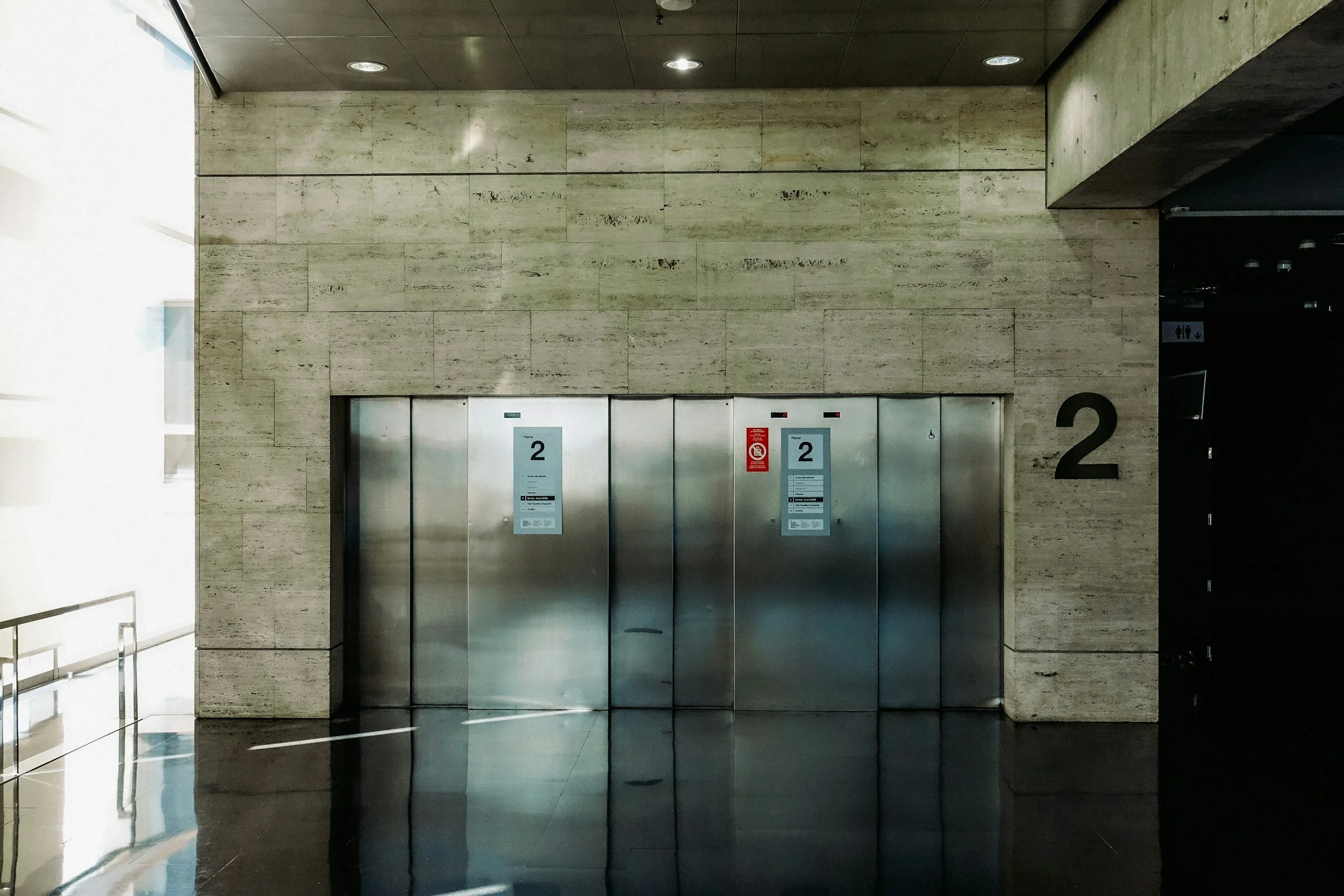 Elevator doors in a building with beige marble walls and black floor. The doors have signs indicating floor 2 and accessibility.