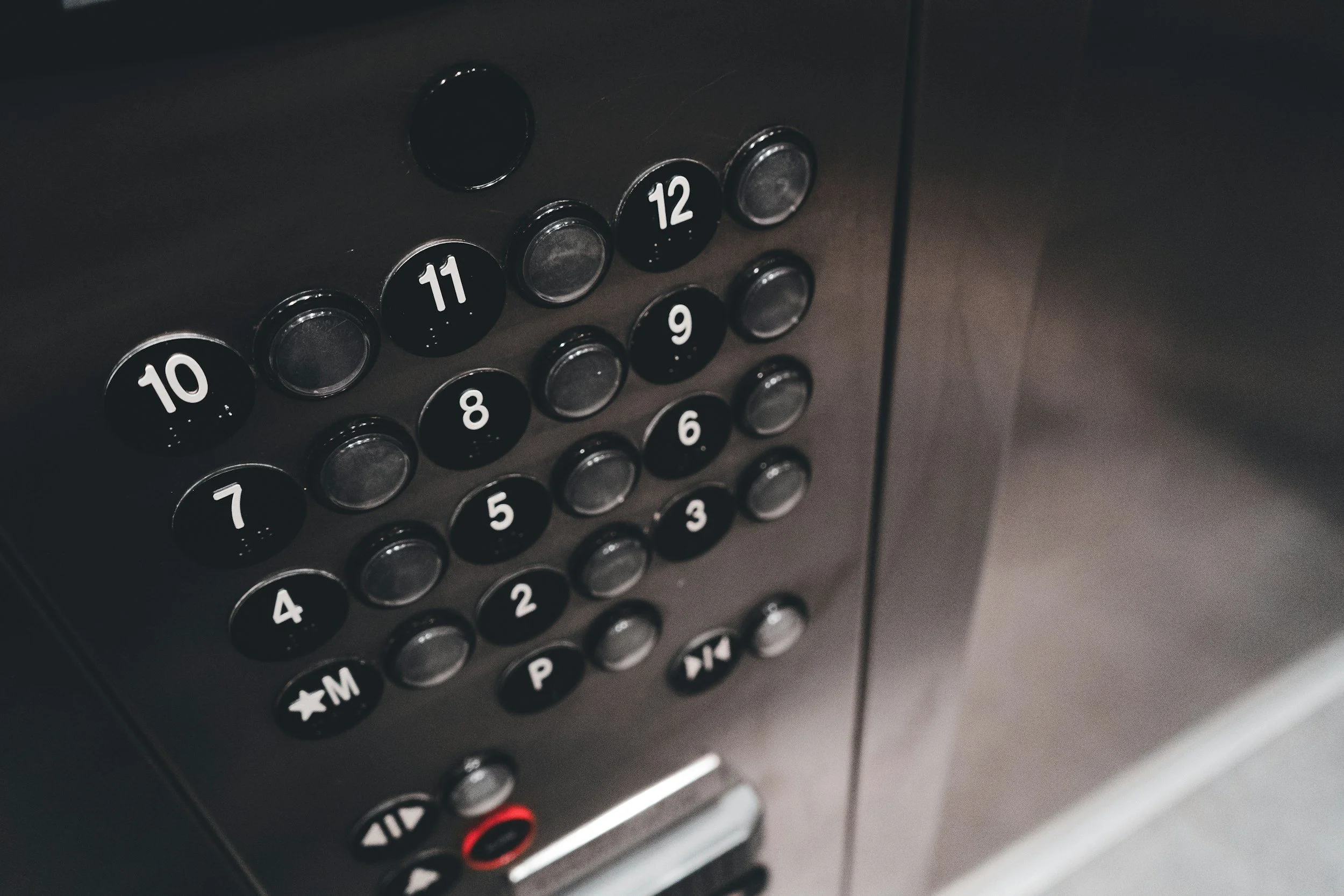 Close-up of elevator control panel with black buttons numbered 1 through 12, including additional function buttons, in a dimly lit elevator.