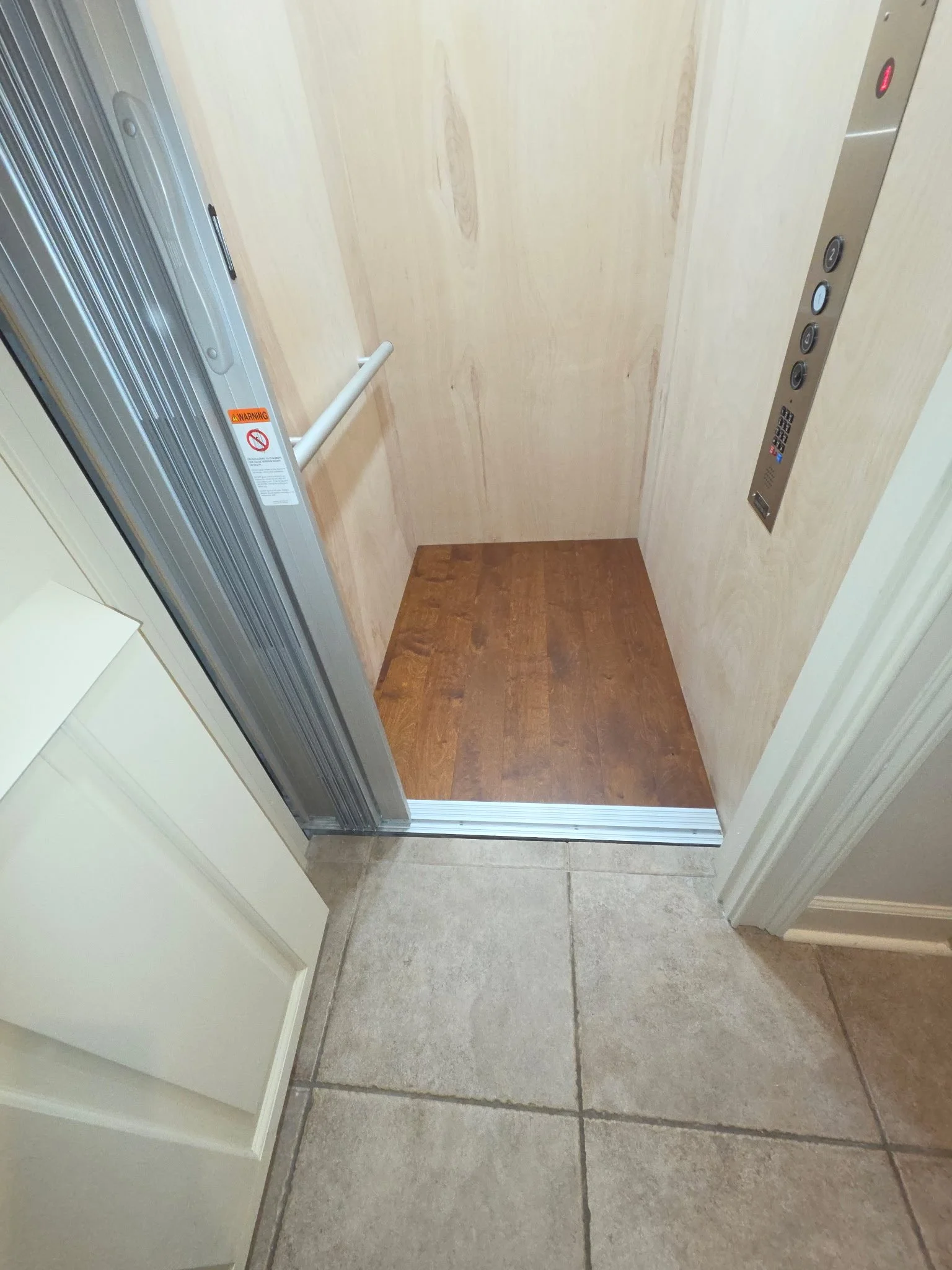 Elevator with open doors, control panel on right wall, wooden floor inside, and tiled entrance floor outside.