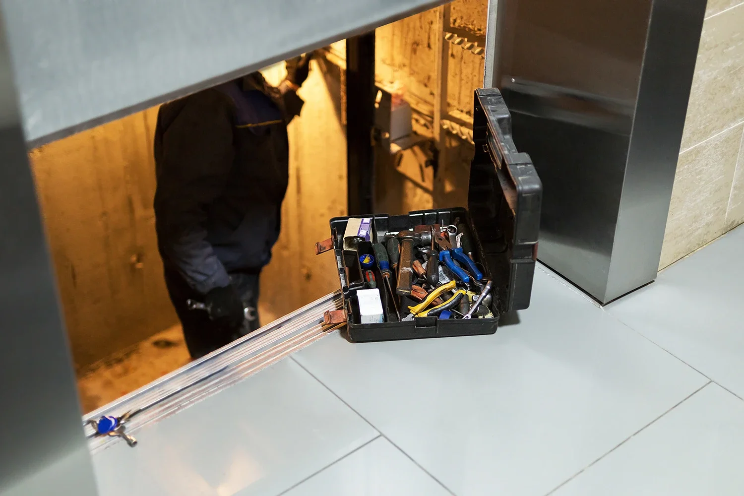 A toolbox filled with various hand tools such as pliers, screwdrivers, and hammers positioned on the floor near a service hatch, with a person partially visible behind the hatch.