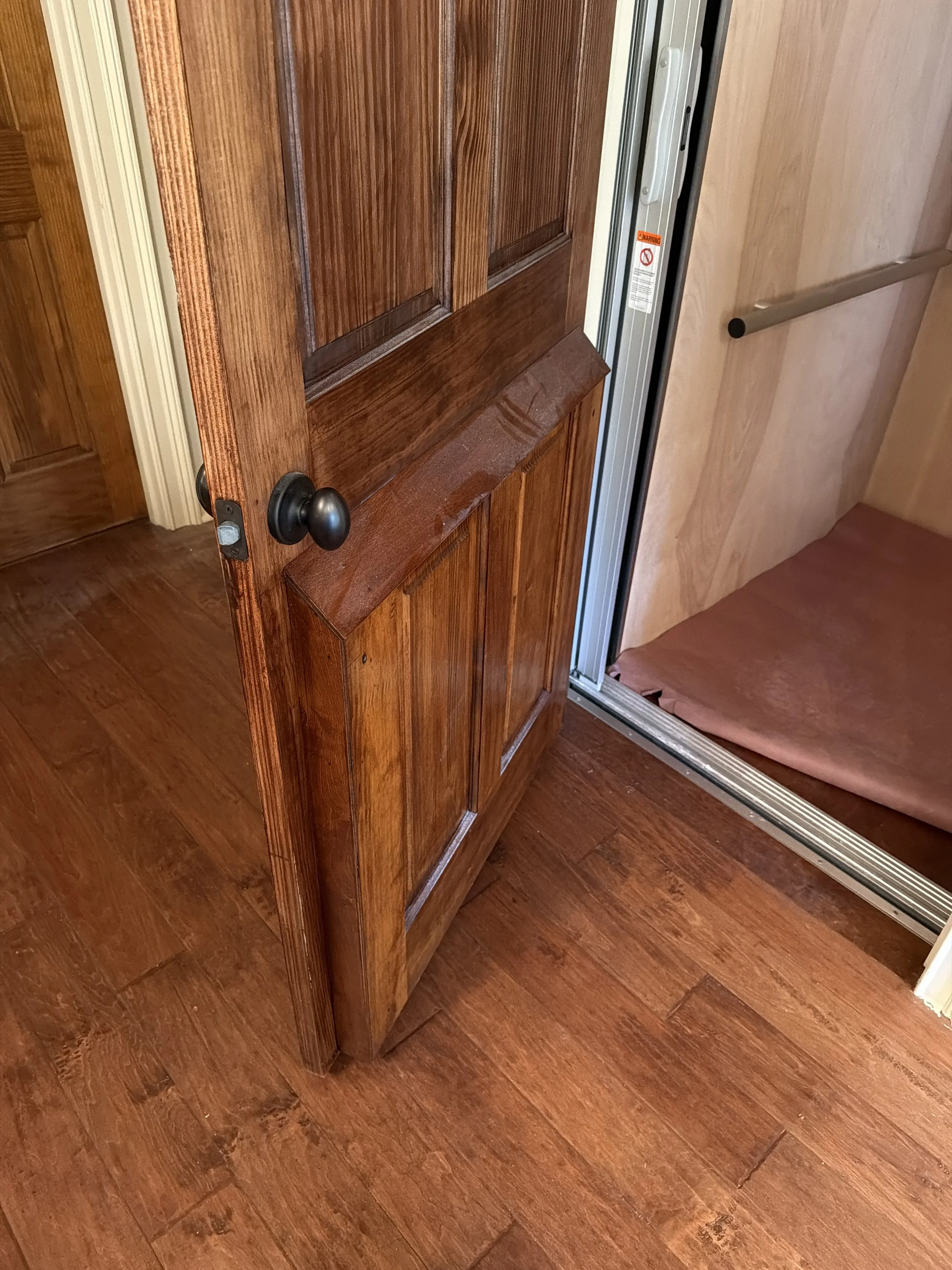 Wooden door with black doorknob open, revealing a partially visible elevator with a brown cushioned floor inside and a metal handrail inside the elevator.