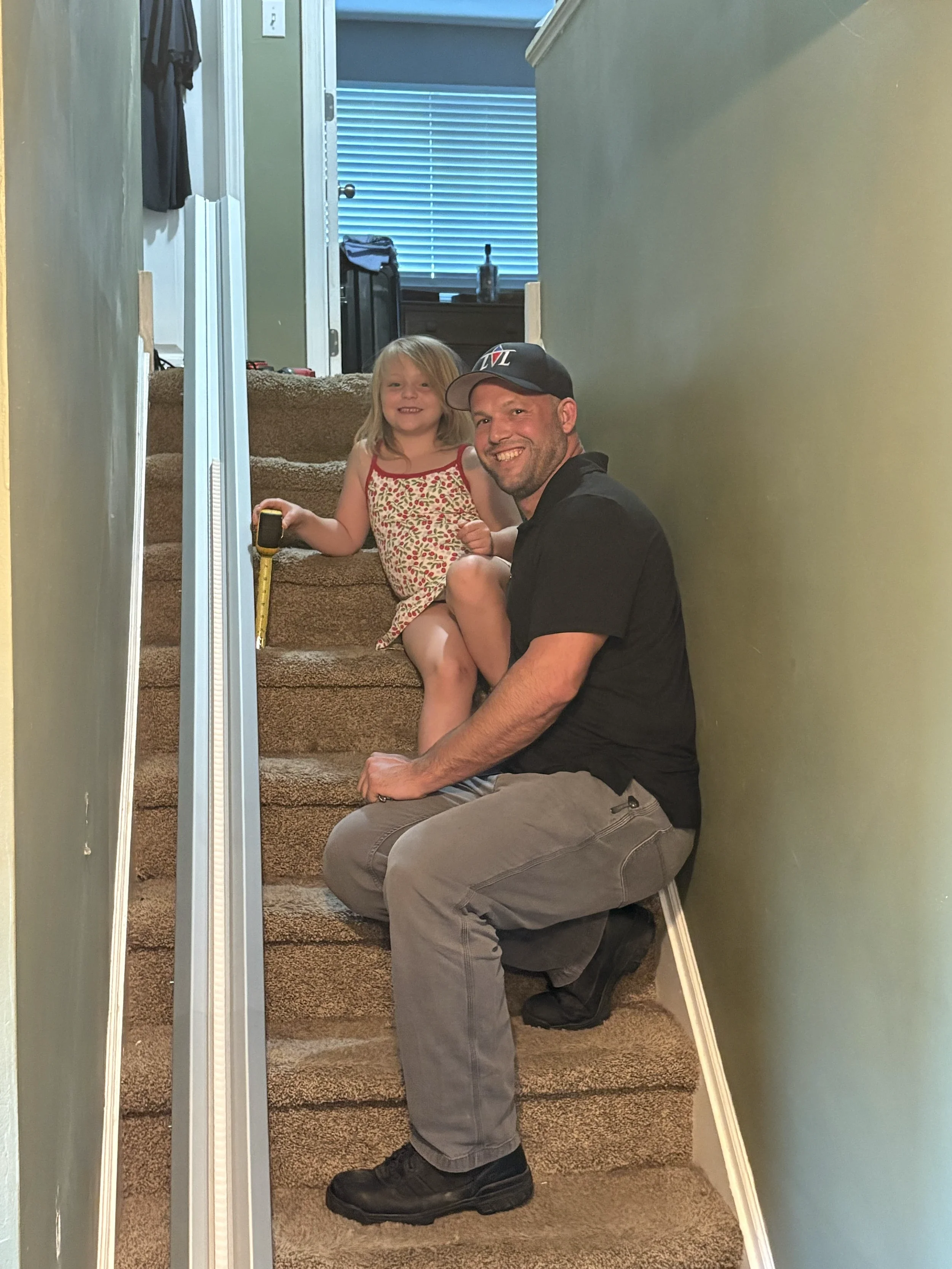 A man and a young girl sitting on a carpeted staircase in a hallway, smiling at the camera. The girl is holding a yellow measuring tape and a remote control, wearing a sleeveless dress with a floral pattern. The man is dressed in a black shirt, gray pants, and black boots, and is wearing a black baseball cap. Behind them, there is a window with closed blinds and a bottle on the windowsill.