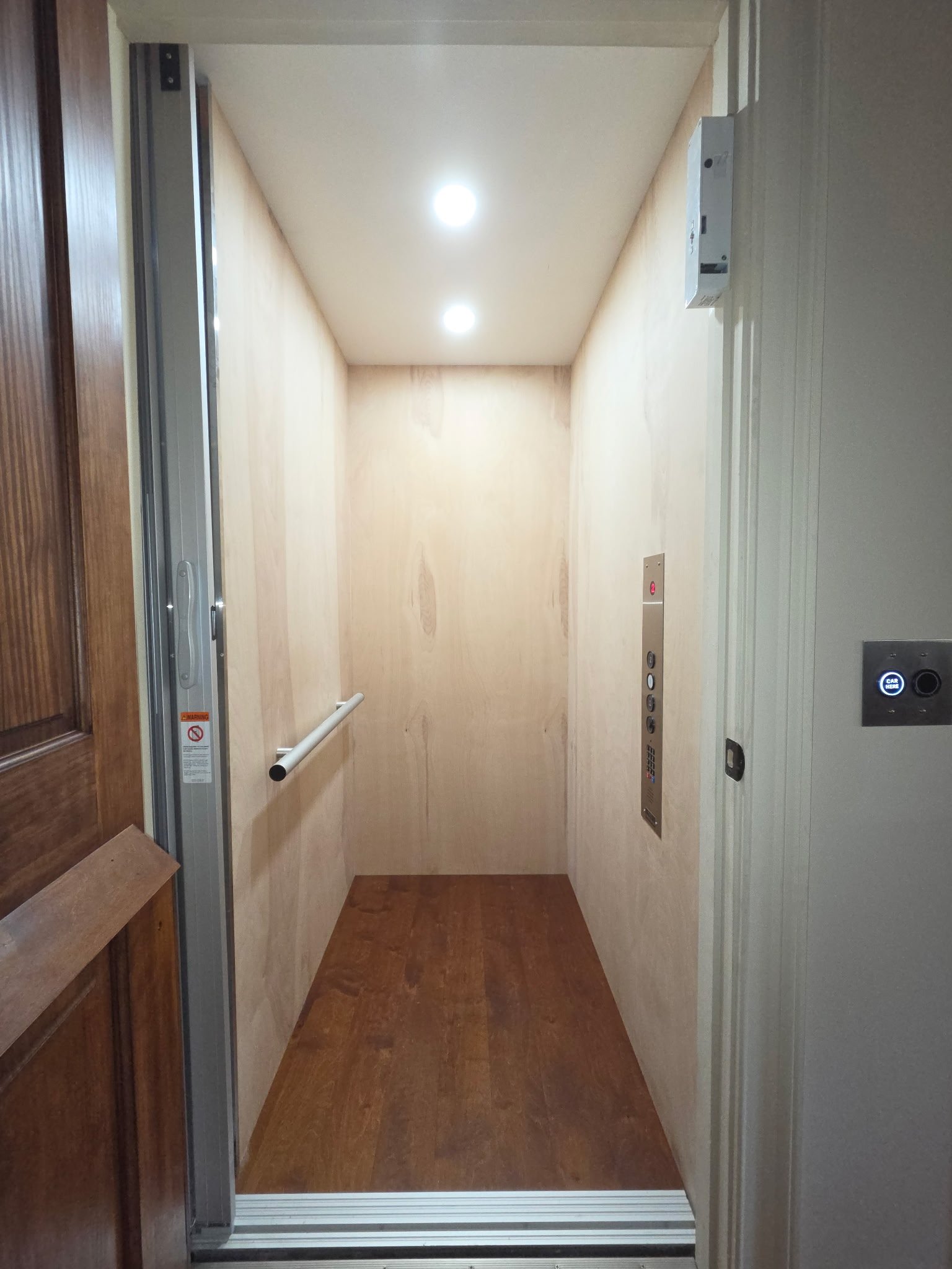 An empty elevator with wooden walls, wood flooring, and a white ceiling with two ceiling lights. The elevator has a handrail on the left wall, control panel with buttons, and a card reader.