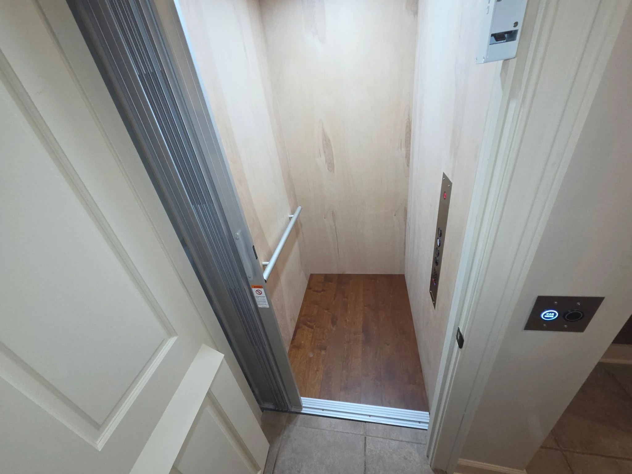 The image shows an open elevator with a wooden floor, metallic door frame, control panel, and buttons, with a small area of wall with decorative molding visible.