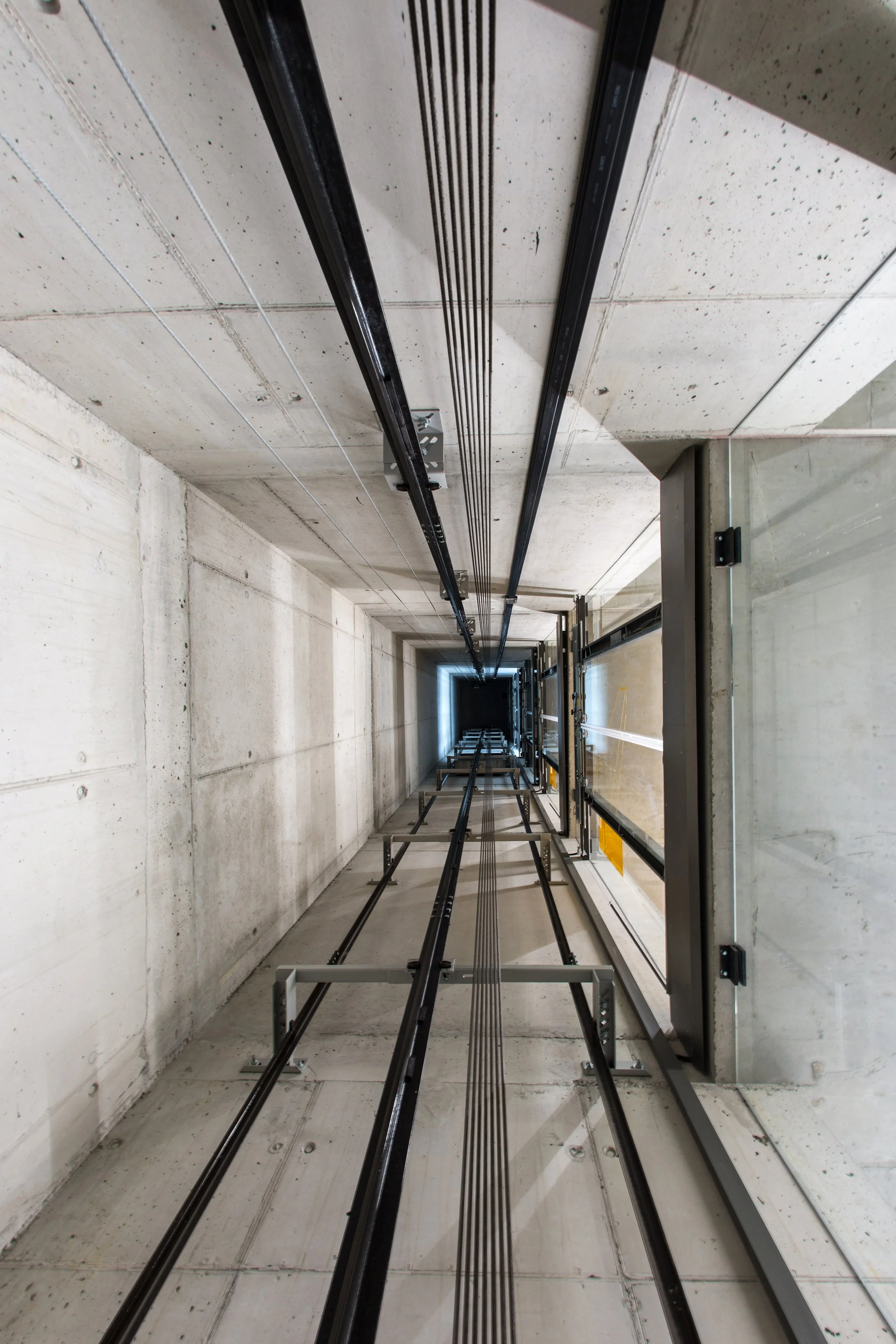 Interior view of an elevator shaft with black cables and metal rails, concrete walls, and a ceiling showing metal supports and wiring.