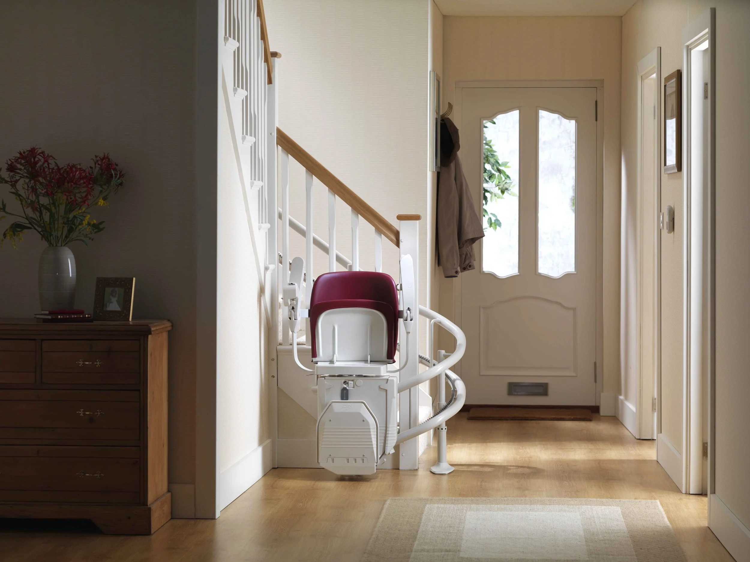 An entrance hallway with a door and hallway walls, showing a stairlift chair attached to the staircase railing, with a coat hanging on a hook near the door, and a wooden dresser with a vase of flowers on top.