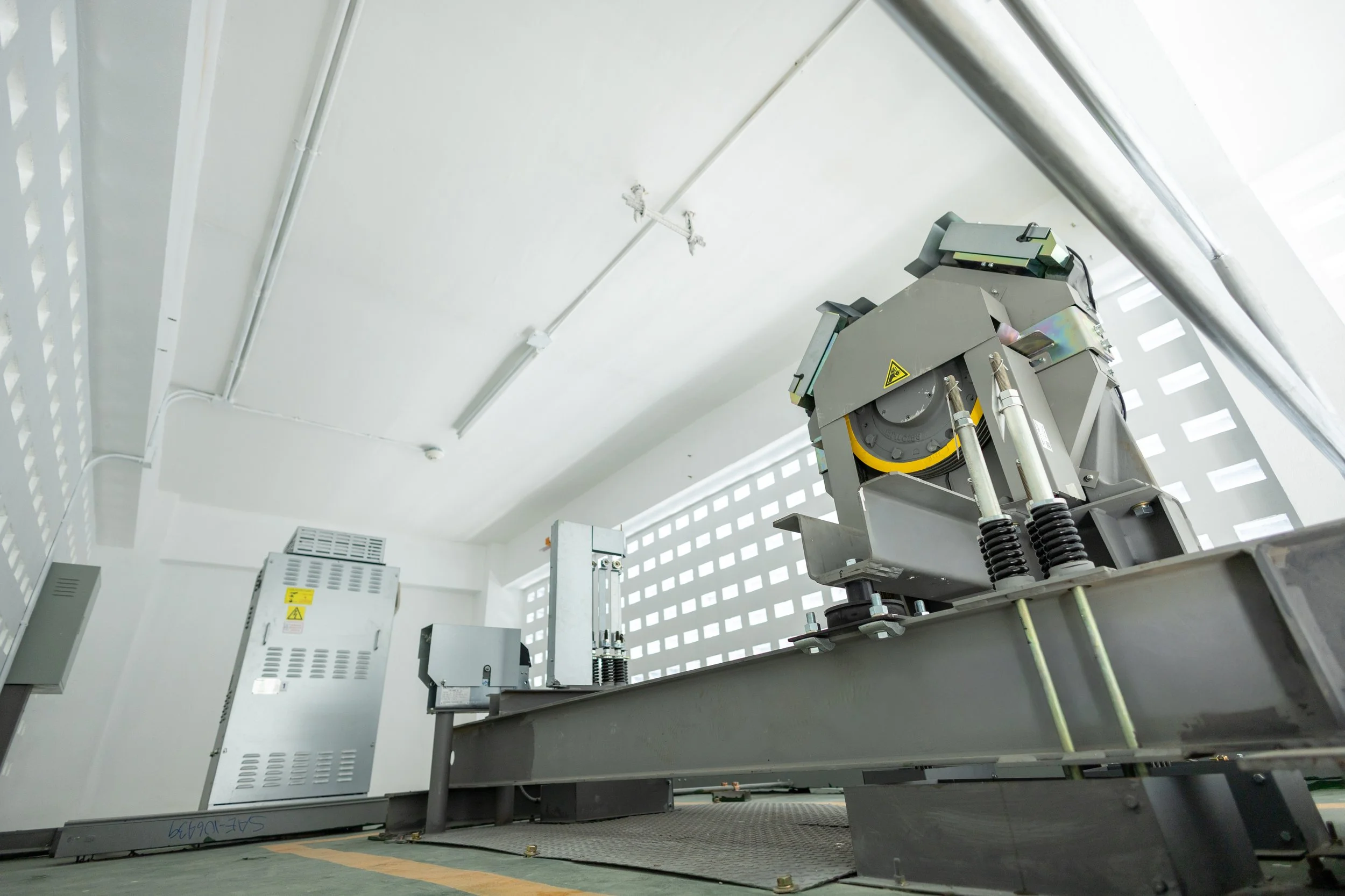 Industrial machinery mounted on a metal platform inside a white room with ventilation panels. The machinery has warning labels and various wires connected.