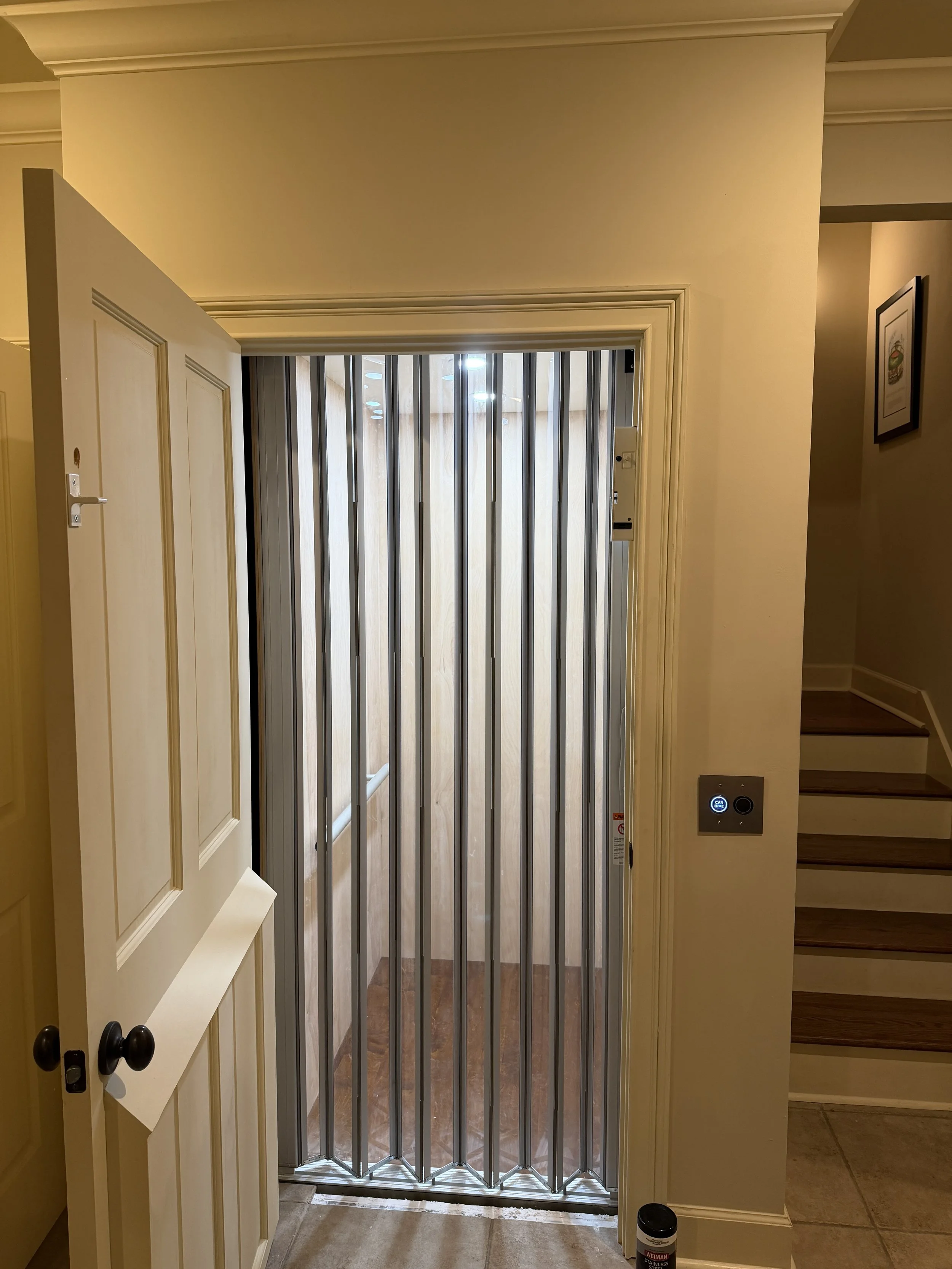 Open elevator door with black knob handles, metal security gate, and wall-mounted control panel, interior of a building with stairs to the right, framed picture on the wall, and tiled flooring.
