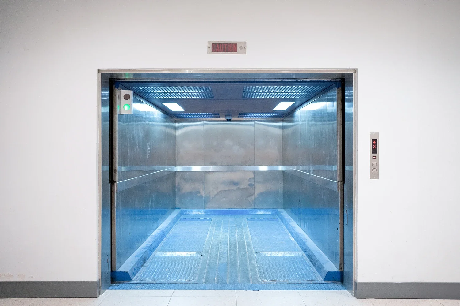 Empty stainless steel elevator with open doors in a white wall corridor.