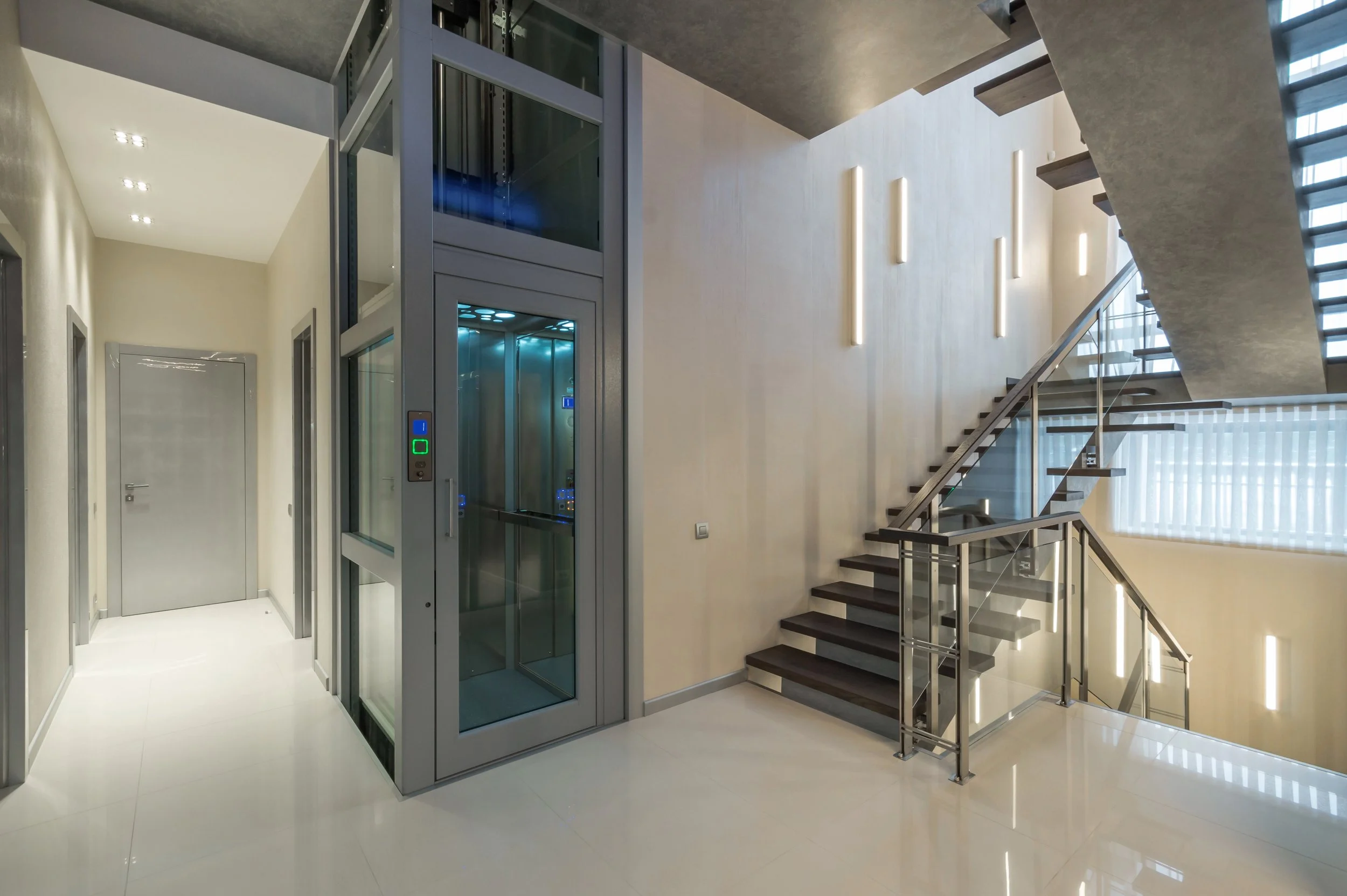 Modern hotel lobby featuring a glass elevator, stairs with metal railings, and contemporary lighting fixtures.