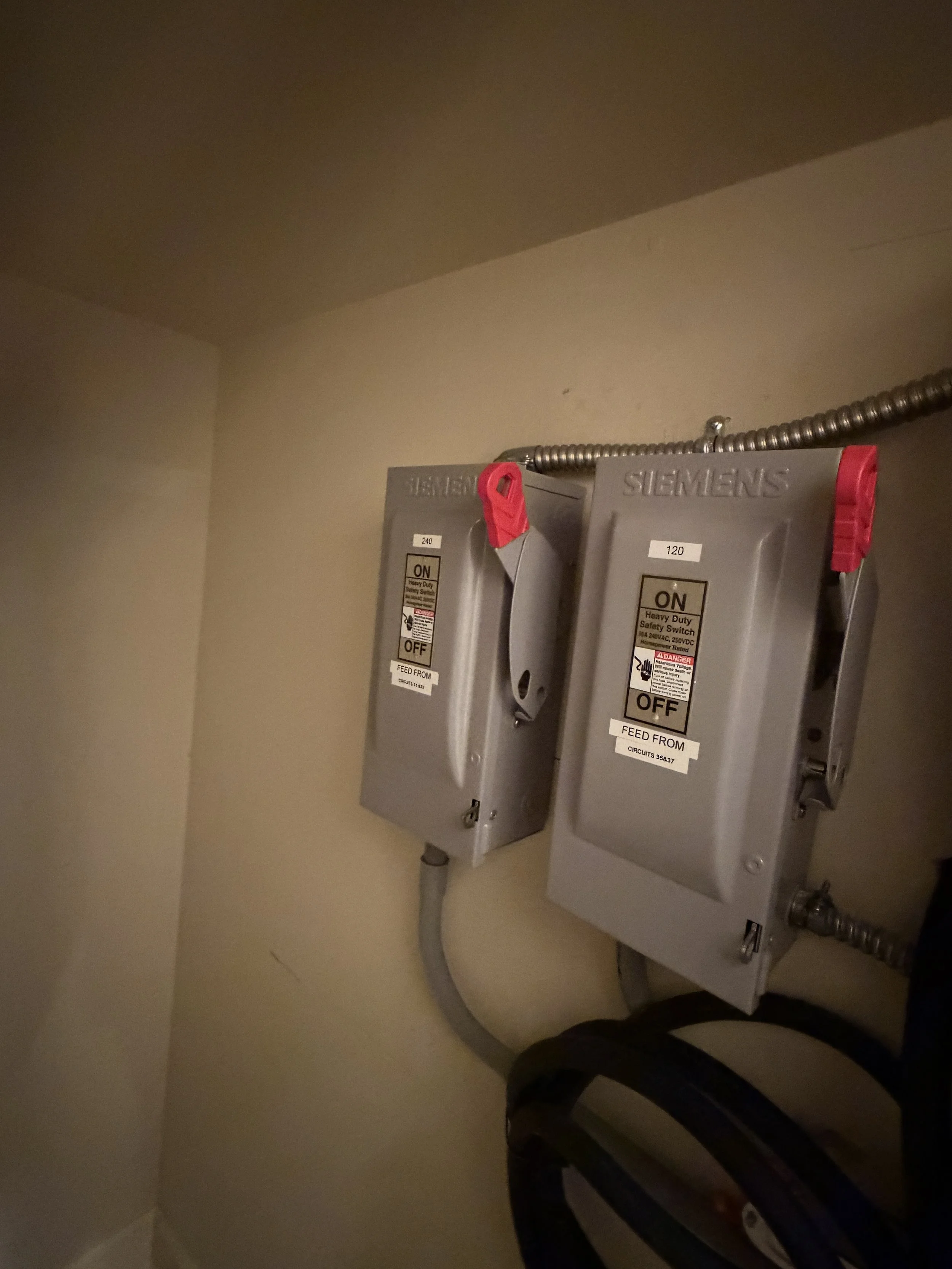 Electrical circuit breakers in a gray metal box mounted on a beige wall with conduit pipes.