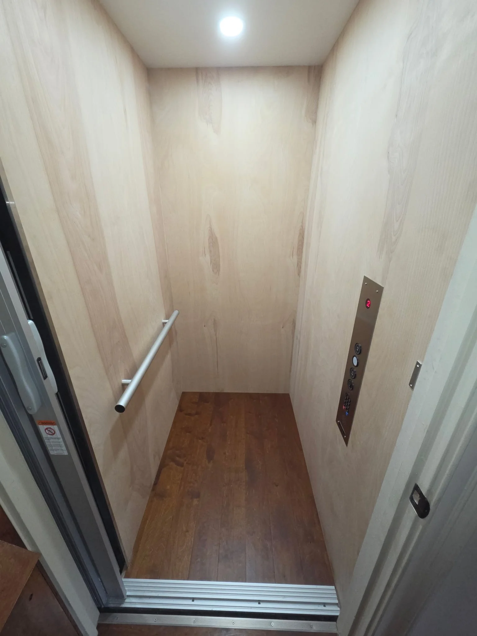 Inside an elevator with wooden panel walls, floor, overhead light, handrail, and control panel with buttons.