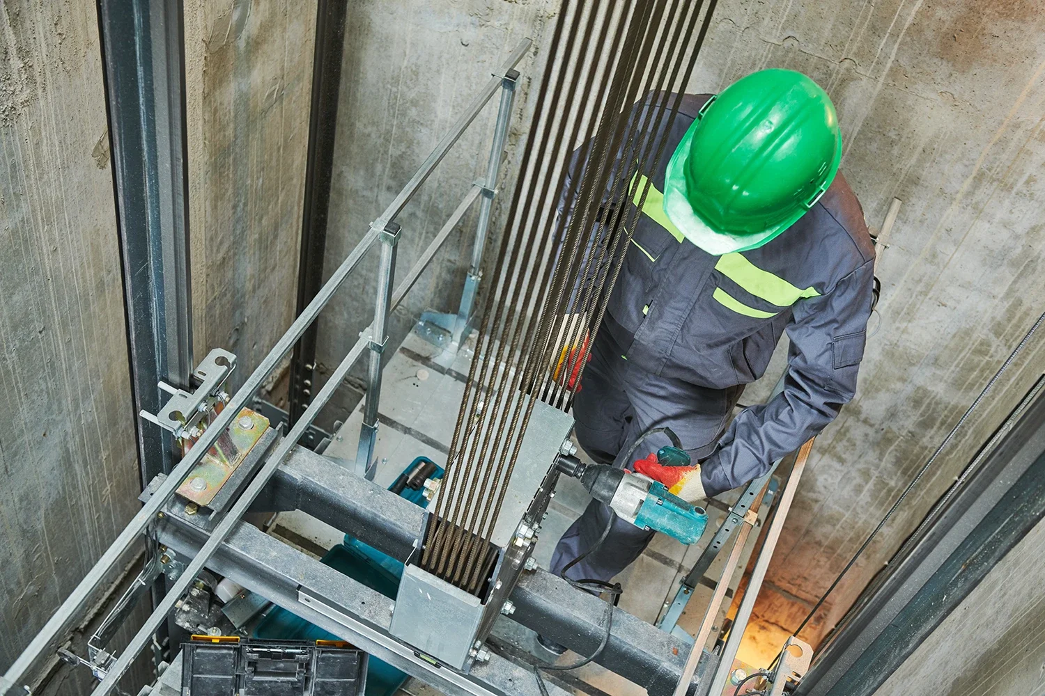 Construction worker wearing a green safety helmet, gray work uniform with reflective stripes, and gloves, operating a power drill while standing in a construction elevator.