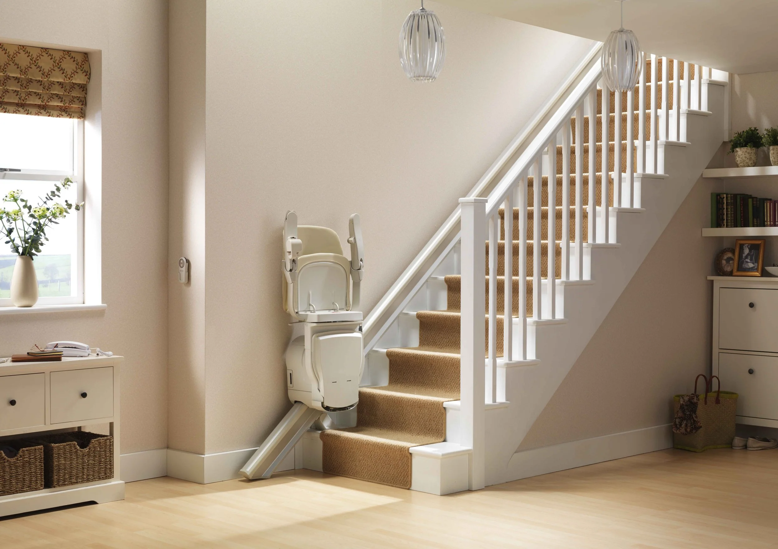 A staircase with beige carpet, white railing, and two hanging light fixtures in a well-lit room. A white high chair is positioned near the stairs, and there are shelves with books and a potted plant on the wall.