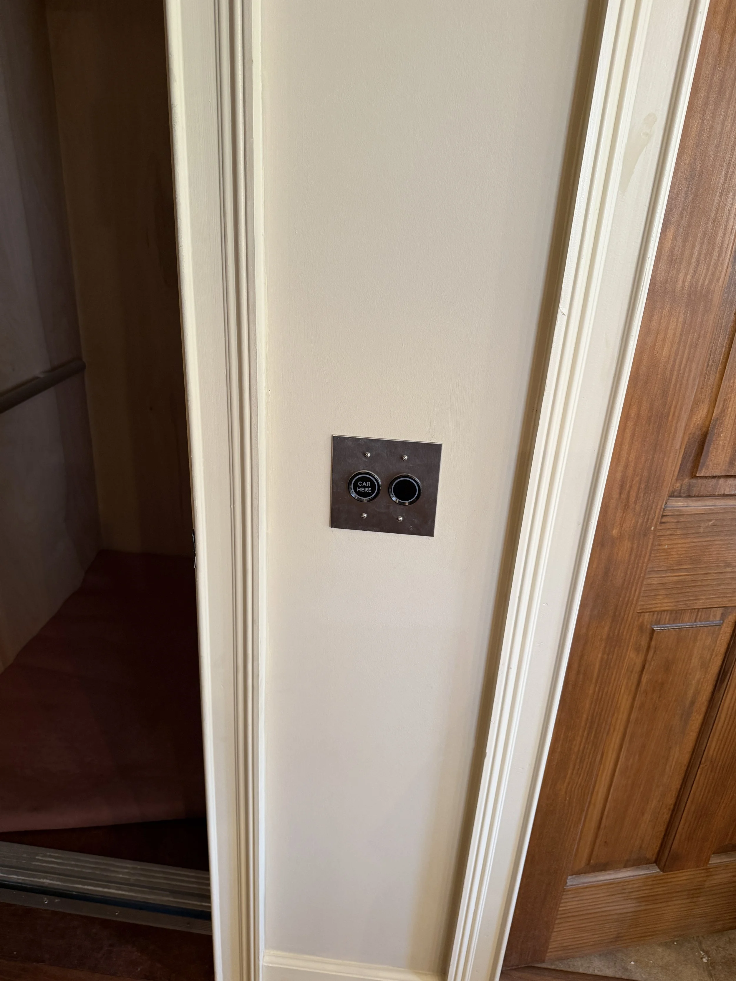 The elevator door is open, revealing a dark interior with wood flooring inside. The elevator control panel on the wall has two buttons, one labeled 'CAR HERE' and the other is blank.