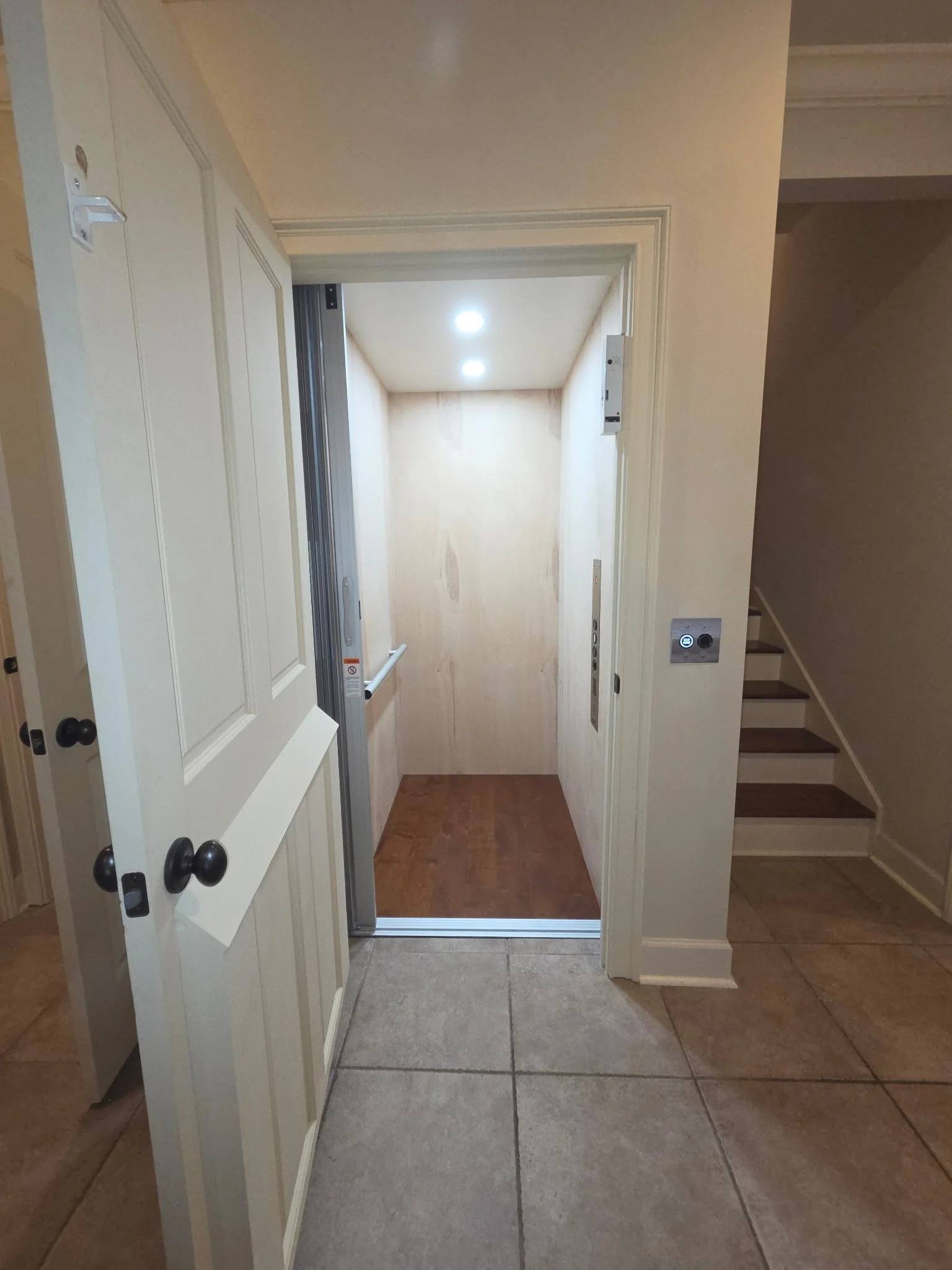 An empty elevator with closed doors, wooden flooring, and beige walls, adjacent to a staircase with wooden steps and a beige wall.