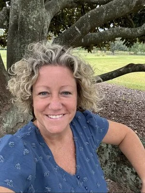 A woman with curly gray hair smiling outdoors, sitting near a large tree in a park.