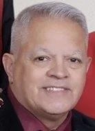 Portrait of an older man with gray hair smiling, wearing a dark blazer and maroon shirt, against a neutral background.