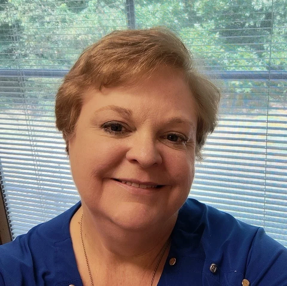 A woman with short, light brown hair smiling, wearing a blue top, sitting in front of a window with blinds and green trees visible outside.