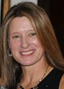 A woman with long, light brown hair, smiling, wearing a black top and earrings.