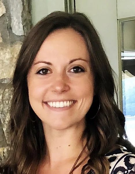 A young woman with long, wavy brown hair, smiling, standing in front of a stone wall and window.