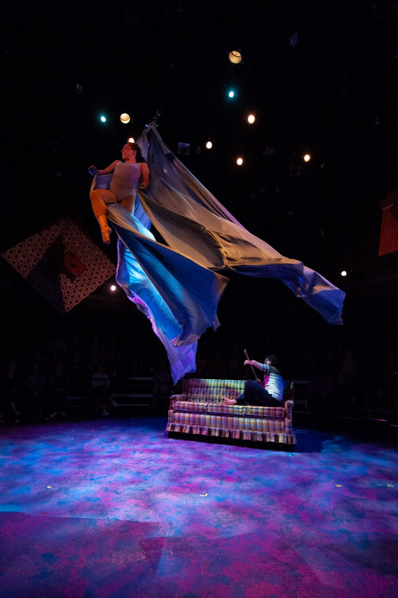 Aerial performer suspended in blue fabric above a stage with a person sitting on a plaid couch, under dramatic colorful lighting. Noah Dach creative director.