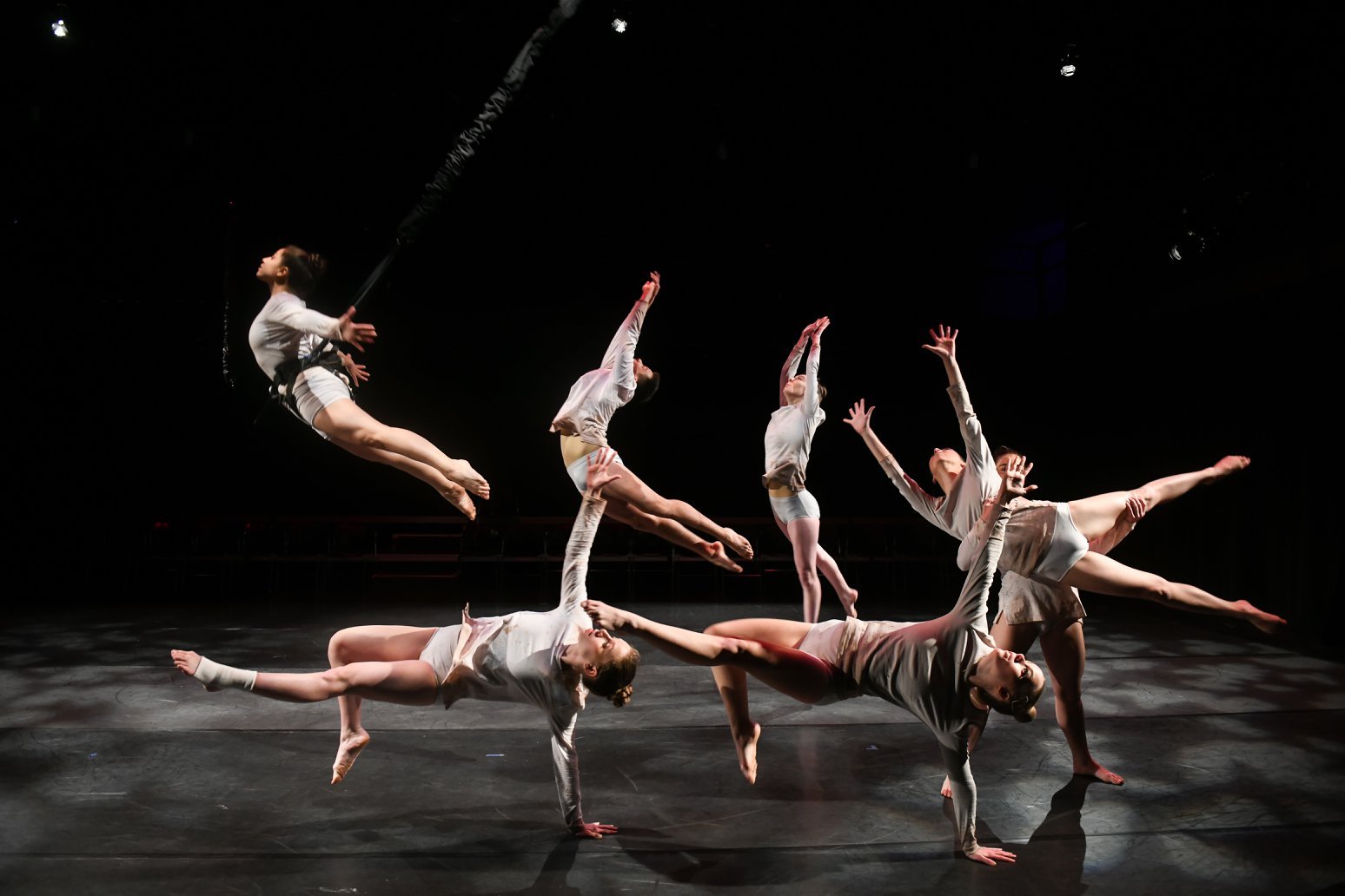 Contemporary dancers performing mid-air on stage with dramatic lighting and shadows, wearing white costumes. Noah Dach creative director.