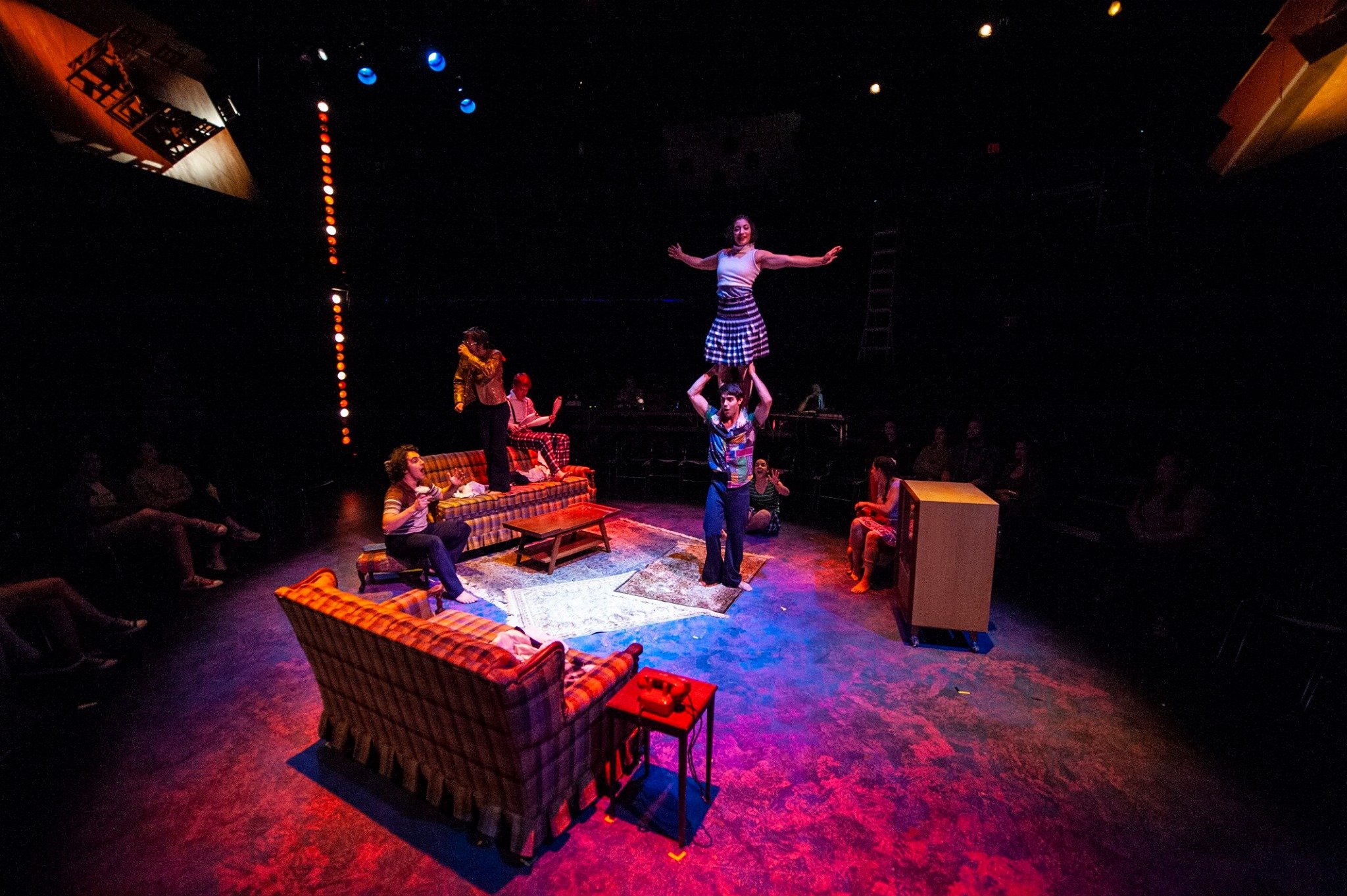 A theater performance featuring a living room set with couches and a small table. Multiple performers are acting and one performer is balancing another on their shoulders. The stage is dimly lit with colorful spotlights and the audience is visible in