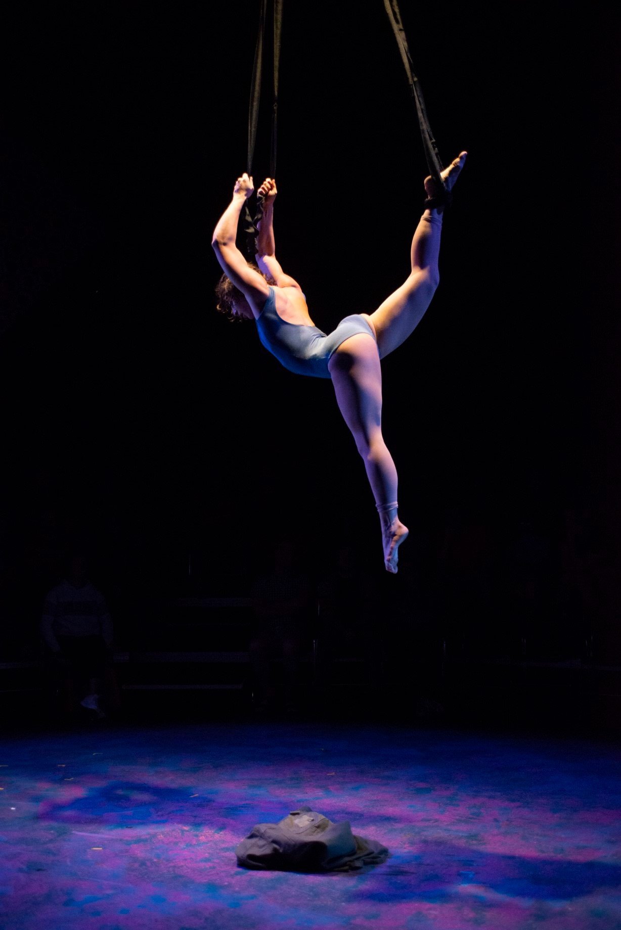 Aerial performer suspended, doing a split in mid-air with a dark background. Noah Dach creative director.