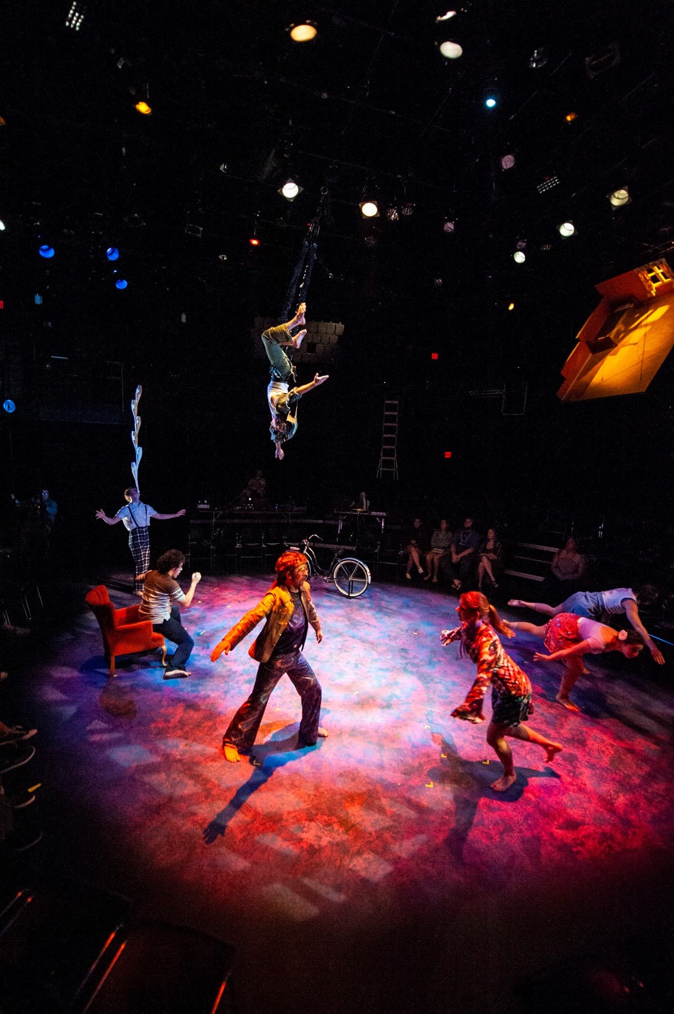 Circus performers in a dimly lit theater; one hanging upside down, others dancing on a colorful stage. Noah Dach creative director.