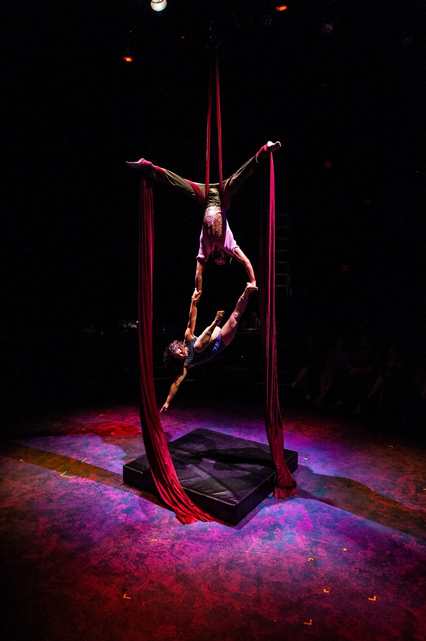 Two aerialists performing on red silks during a circus show, with one upside down holding the other below, on a dimly lit stage. Noah Dach creative director.