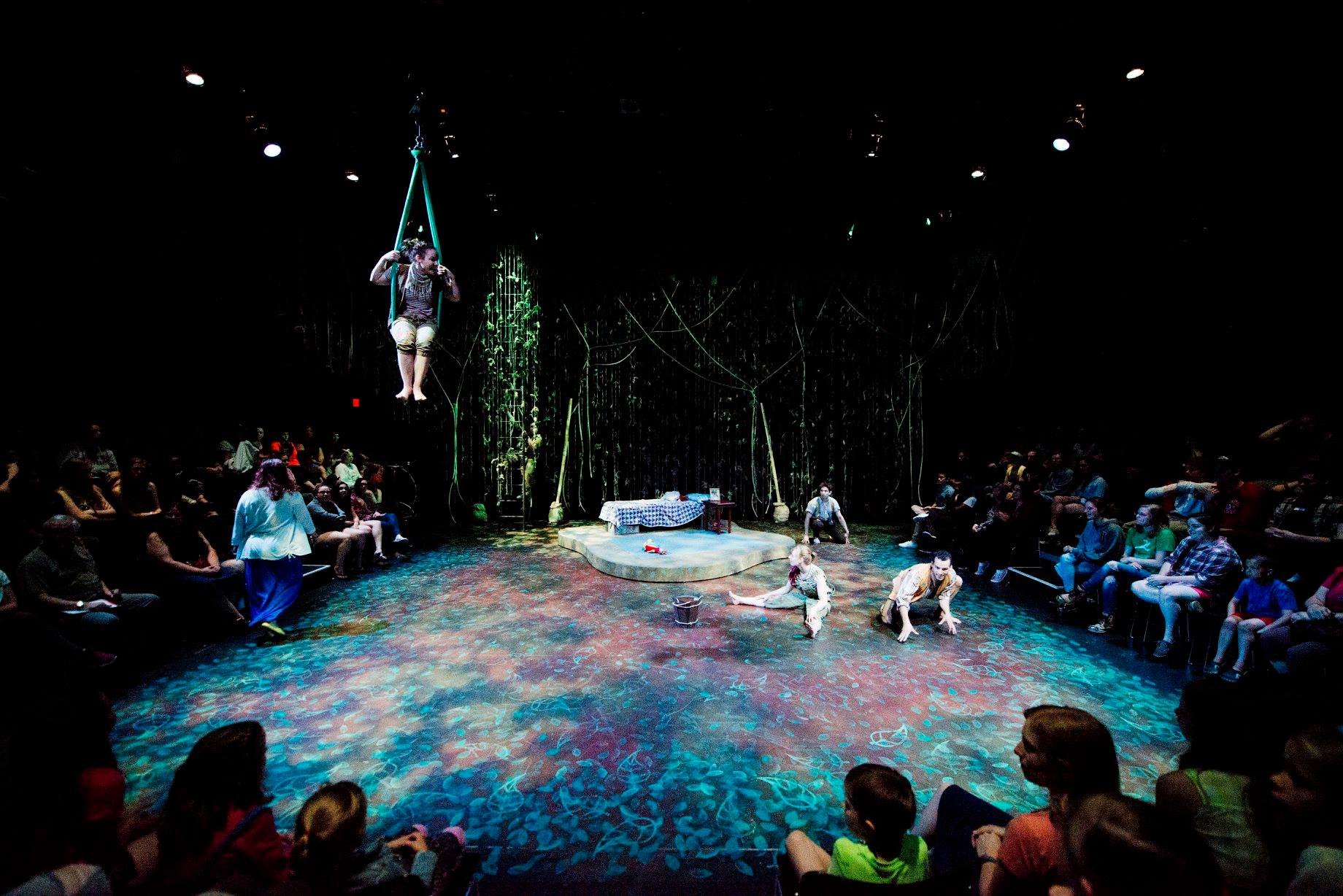 Theatrical performance in a circular stage setting with an audience surrounding. One performer is suspended from above, while others act on stage. The floor is painted with vivid colors, and stage lighting creates a dramatic effect. Noah Dach creativ