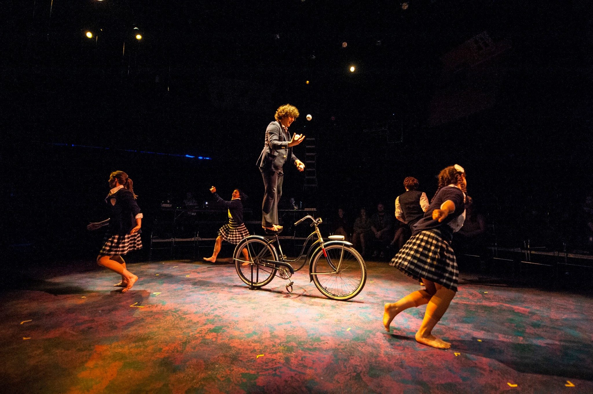 Circus performer balancing on a bicycle while juggling balls, surrounded by dancers in checkered skirts in a dimly lit stage environment. Noah Dach creative director.