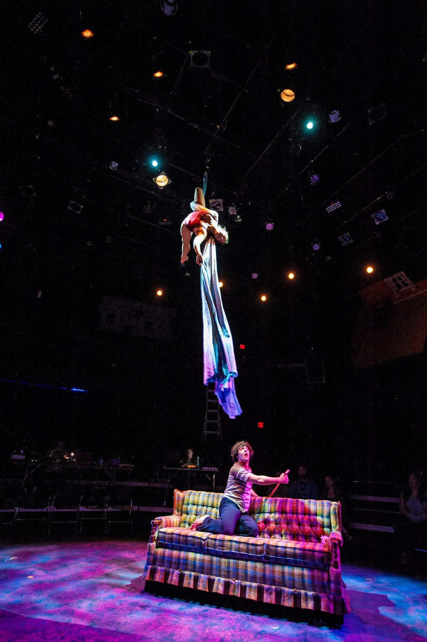 Performer on aerial silks above a colorful couch on stage with dramatic lighting. Noah Dach creative director.