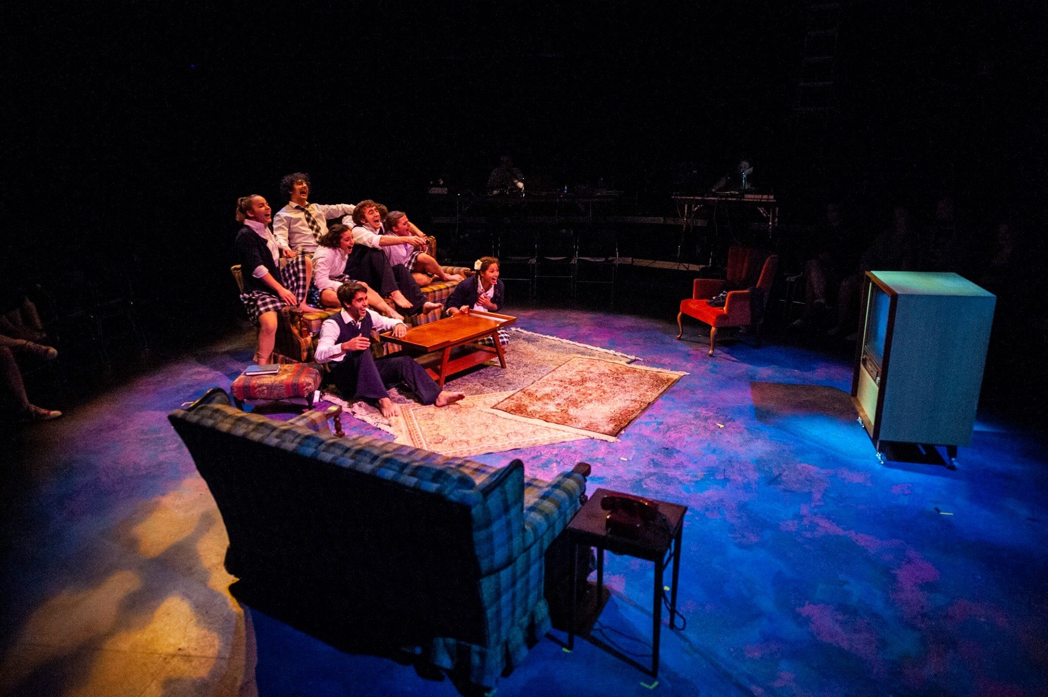 Theatrical stage set with actors on a couch, vintage TV, armchair, and coffee table on a rug. Noah Dach creative director.